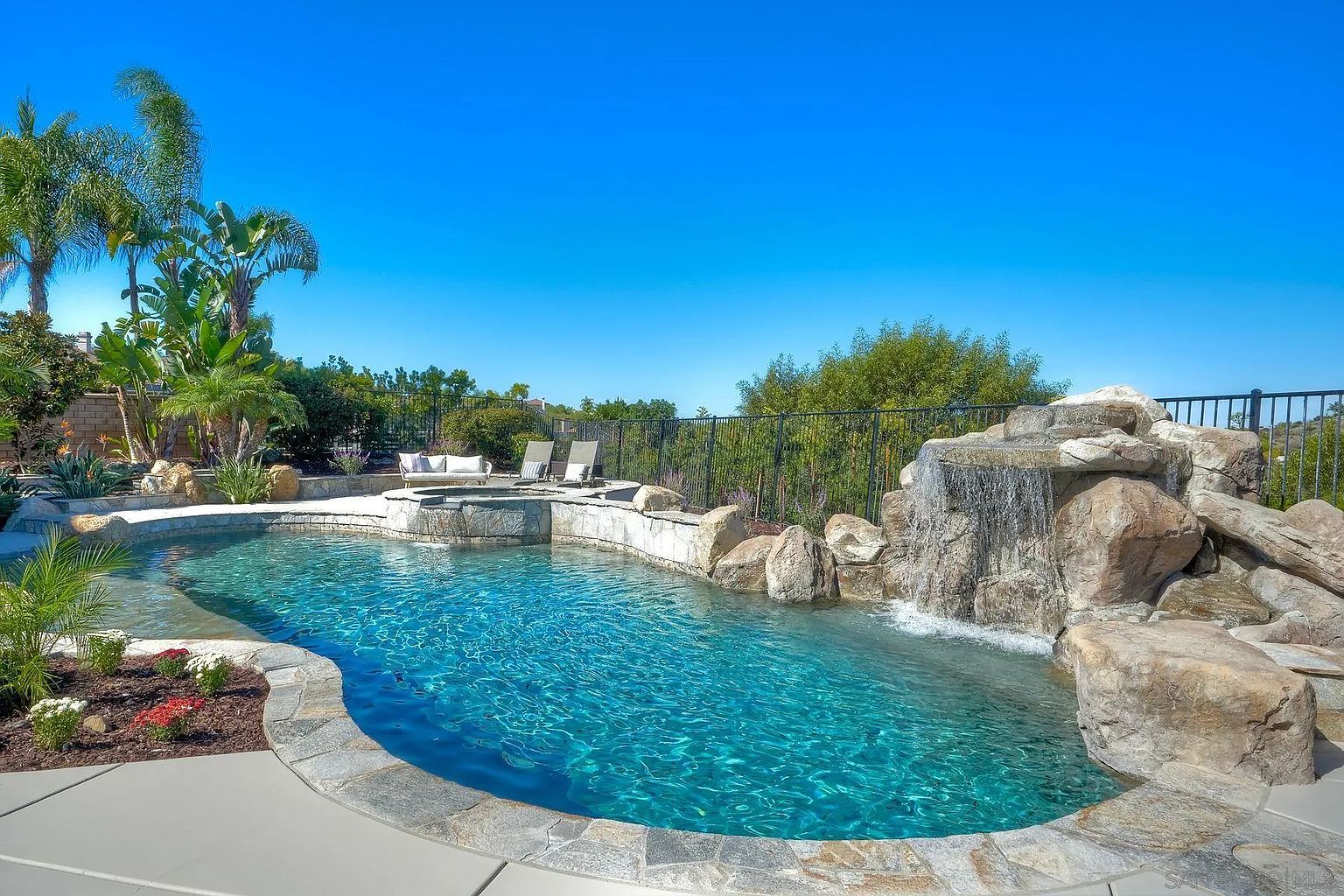 This image showcases a luxurious backyard pool and spa area. The pool features a natural stone waterfall and is surrounded by lush landscaping, including palm trees and flowering plants. Lounge chairs and a seating area are situated near the spa, creating an inviting outdoor living space perfect for relaxation and entertainment.