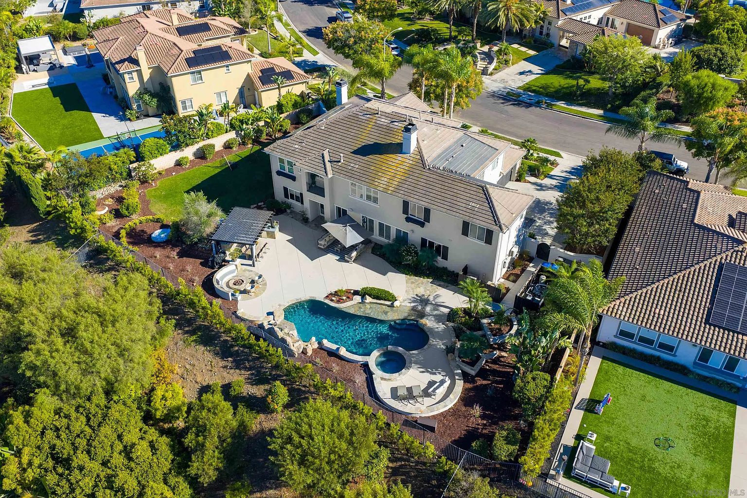 This aerial shot showcases a luxurious home with a stunning backyard oasis. The focal point is a beautifully designed pool and spa area, complemented by a spacious patio with a pergola and fire pit. Lush landscaping surrounds the property, enhancing its privacy and curb appeal.