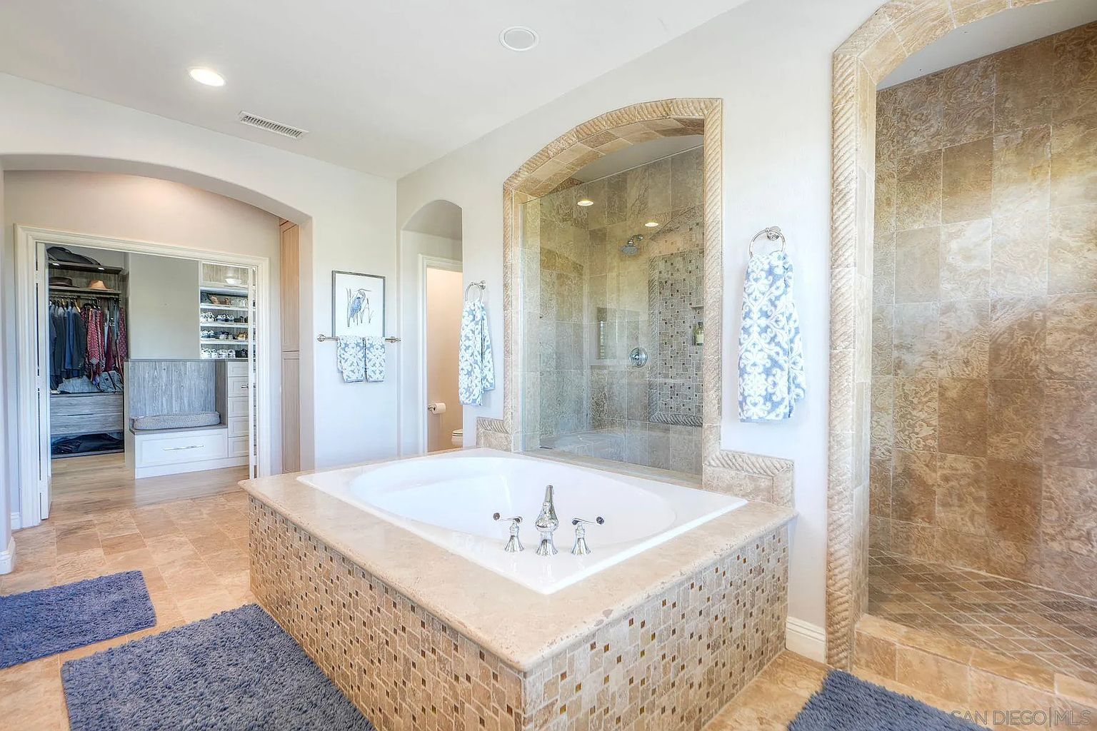This is a luxurious primary bathroom featuring a large soaking tub with mosaic tile surround, a glass-enclosed shower with travertine tile, and an arched doorway leading to a walk-in closet. The color palette is neutral and calming, creating a spa-like atmosphere. The bathroom is spacious and well-lit, with high-end fixtures and finishes.