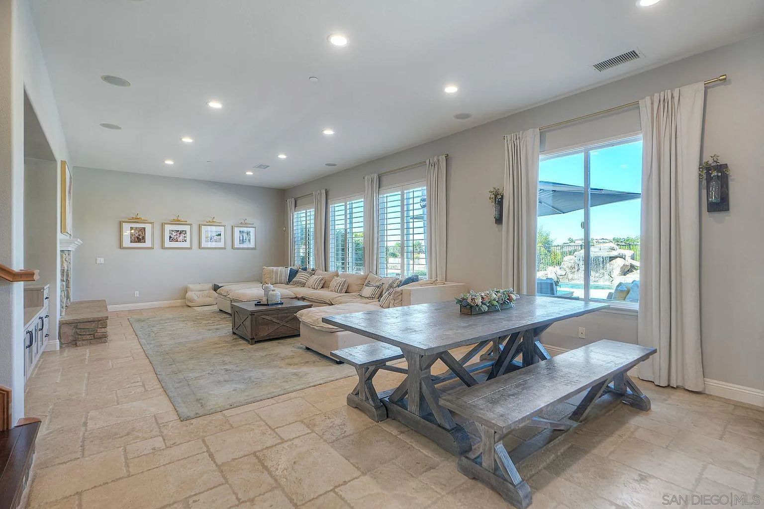 This is an interior shot of a spacious living room featuring a large sectional sofa, a rustic coffee table, and a dining table with benches. The room is well-lit with recessed lighting and natural light from the windows, which are adorned with curtains. The flooring is tiled, and there is a large area rug under the sofa and coffee table, creating a cozy and inviting atmosphere.