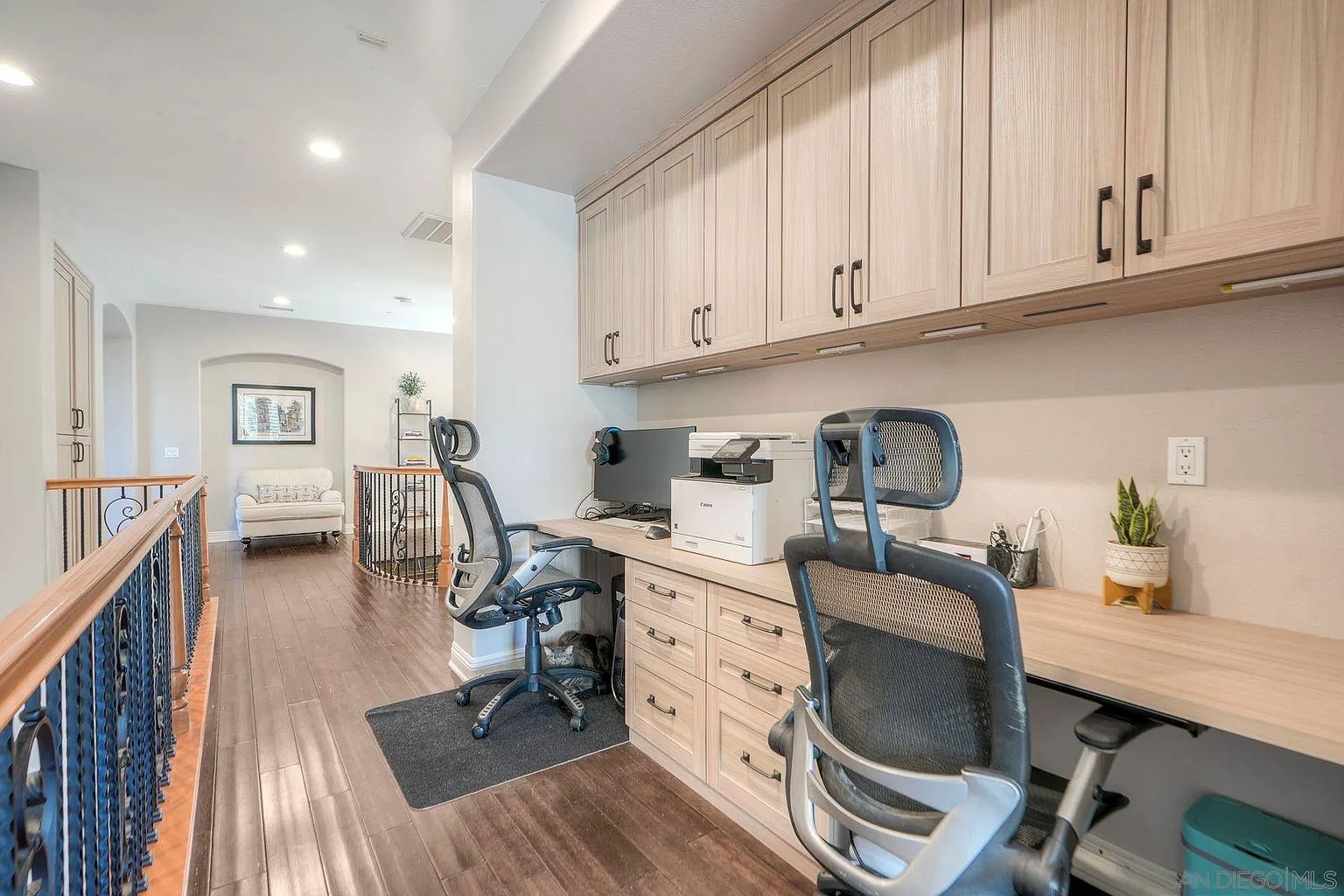 This is an interior shot of a home office space. The office features built-in cabinetry above a long desk, providing ample storage. Two office chairs are visible, suggesting a shared workspace, and the room opens onto a hallway with a decorative railing, creating an open and airy feel.