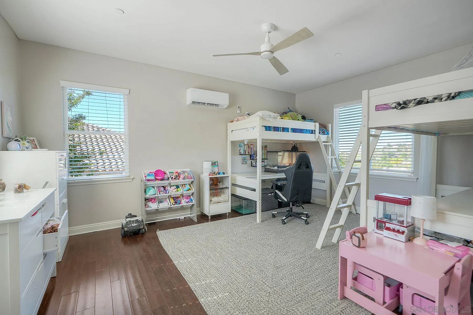 This is a well-organized and inviting children's bedroom featuring a bunk bed with a built-in desk underneath. The room is decorated in neutral tones with pops of color from toys and accessories. A large window provides natural light, and the hardwood floors add warmth to the space. The room also features a pink table and chair set, a toy organizer, and a ceiling fan.