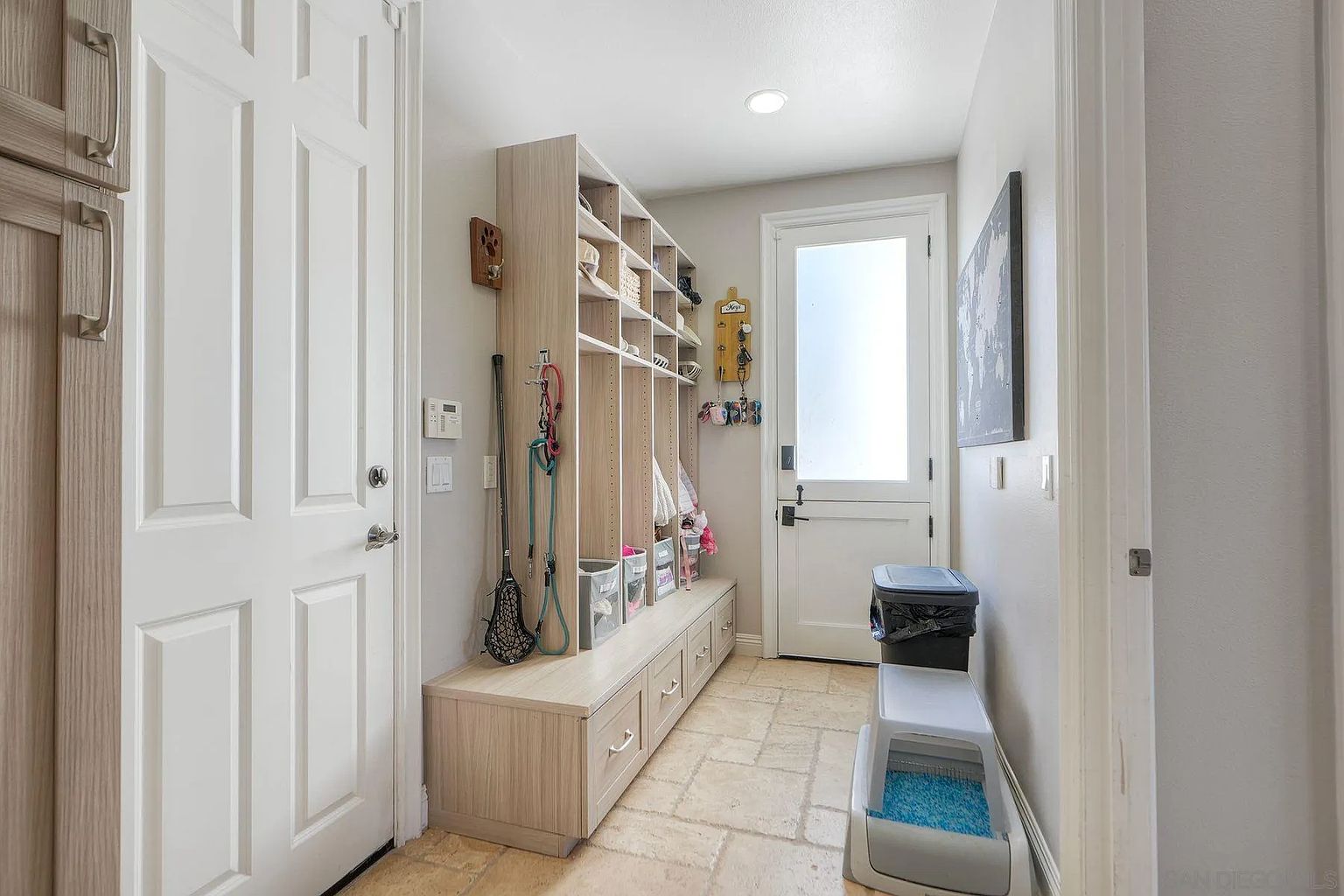 This interior shot showcases a well-organized hallway or mudroom area. The space features a custom built-in storage unit with open shelving, drawers, and hooks for organizing items. A nearby door leads to the outside, and a litter box is placed near the wall, suggesting a pet-friendly home.