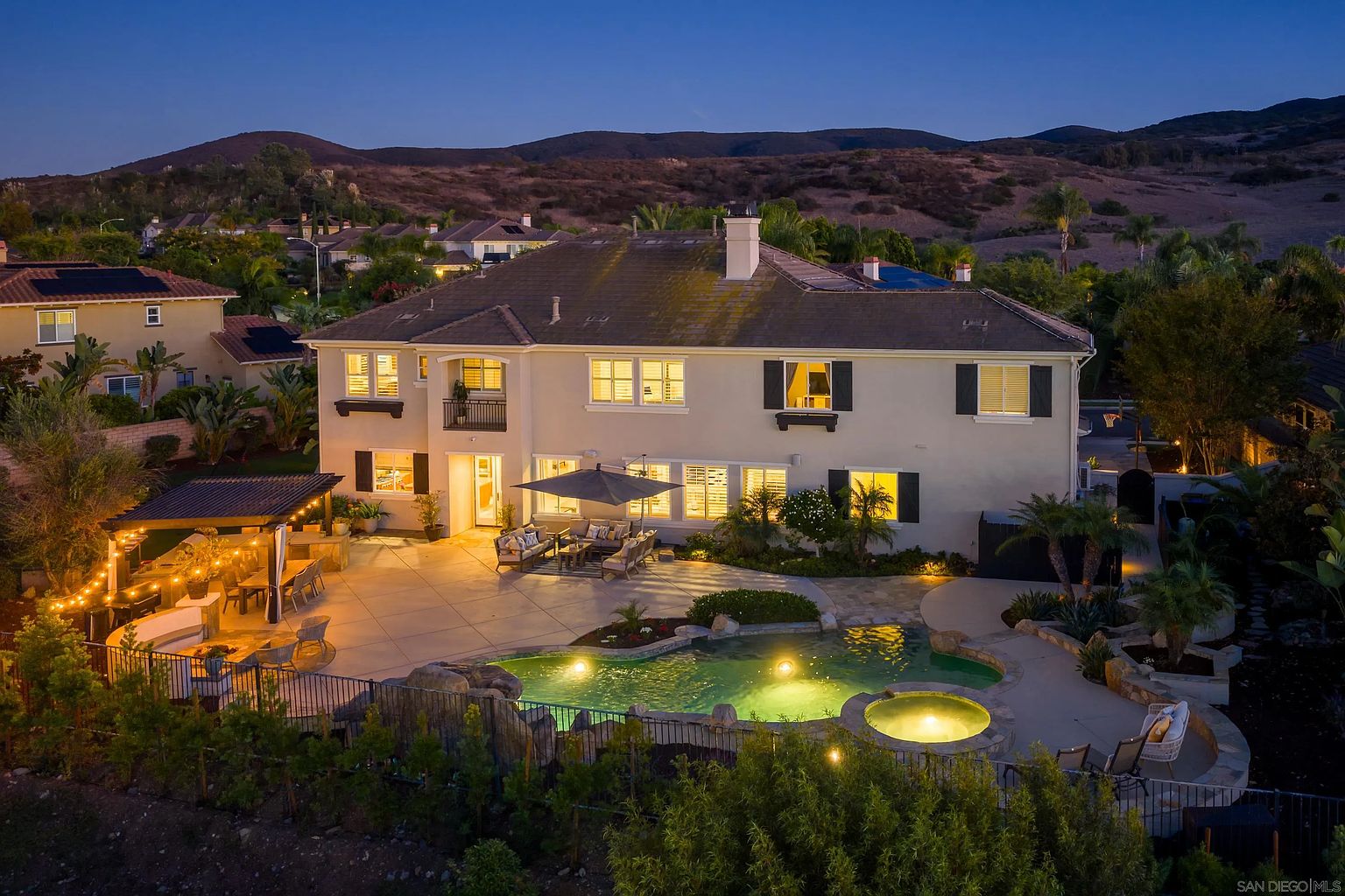 This is a stunning rear view of a two-story home, showcasing a luxurious backyard oasis. The focal point is a beautifully lit pool and spa area, complemented by an outdoor dining space under a pergola adorned with string lights. The well-manicured landscaping and the backdrop of rolling hills create a serene and private atmosphere, perfect for relaxation and entertainment.