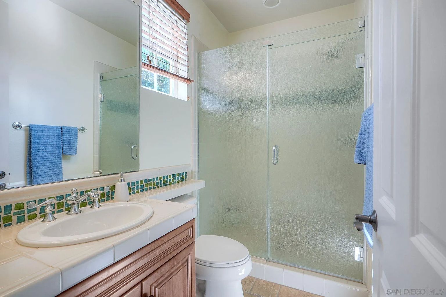 This is a well-lit bathroom featuring a vanity with a white sink and tiled countertop, a wooden cabinet, and a large mirror. A shower with frosted glass doors is visible, along with a toilet. The bathroom has a clean and functional design, suitable for a guest or secondary bathroom.