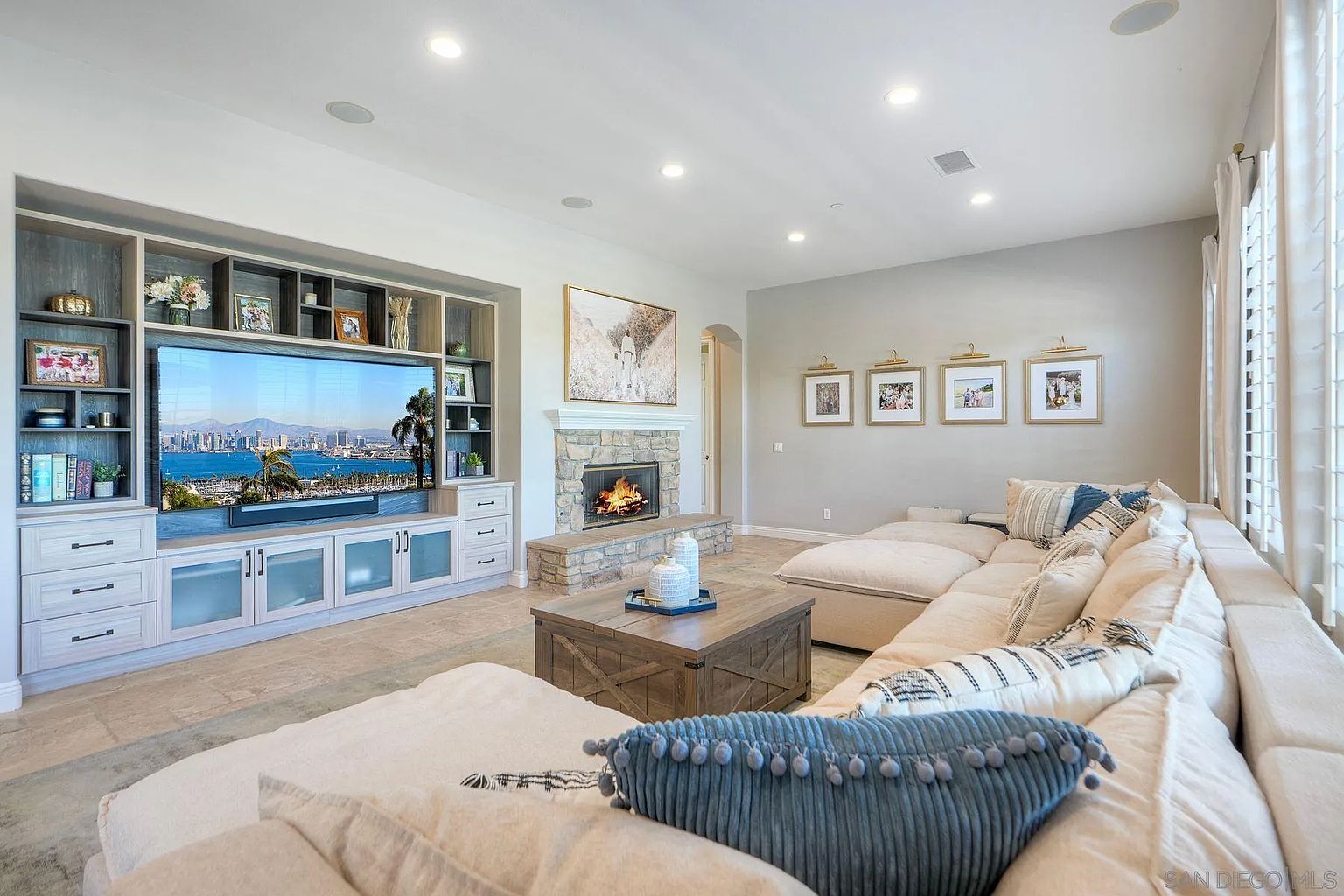 This is an interior shot of a living room featuring a large sectional sofa with decorative pillows, a wooden coffee table, and a stone fireplace. A built-in entertainment center houses a large television. The room is well-lit with recessed lighting and natural light, creating a warm and inviting atmosphere.