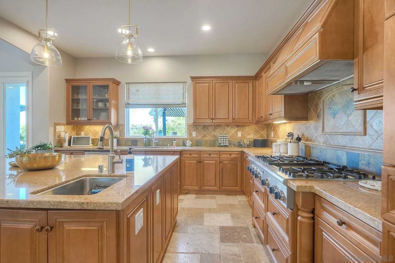 This is a well-lit kitchen featuring custom wood cabinetry, granite countertops, and stainless steel appliances. The kitchen island includes a sink and ample counter space, while a gas range and decorative backsplash add to the room's appeal. The flooring is tiled, and pendant lights hang above the island, creating a warm and inviting atmosphere.