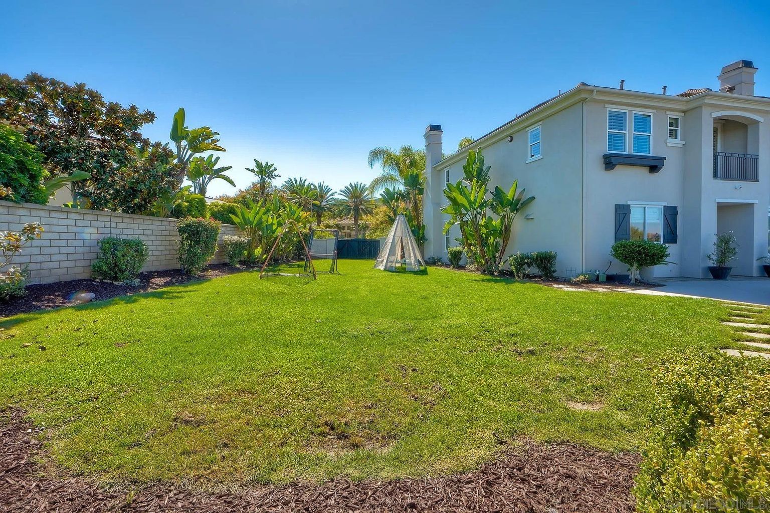 This is a view of the backyard featuring a well-maintained lawn, mature trees, and landscaping. A two-story house is visible on the right side of the frame. A swing set and a teepee-like structure are present in the yard, suggesting a family-friendly environment.