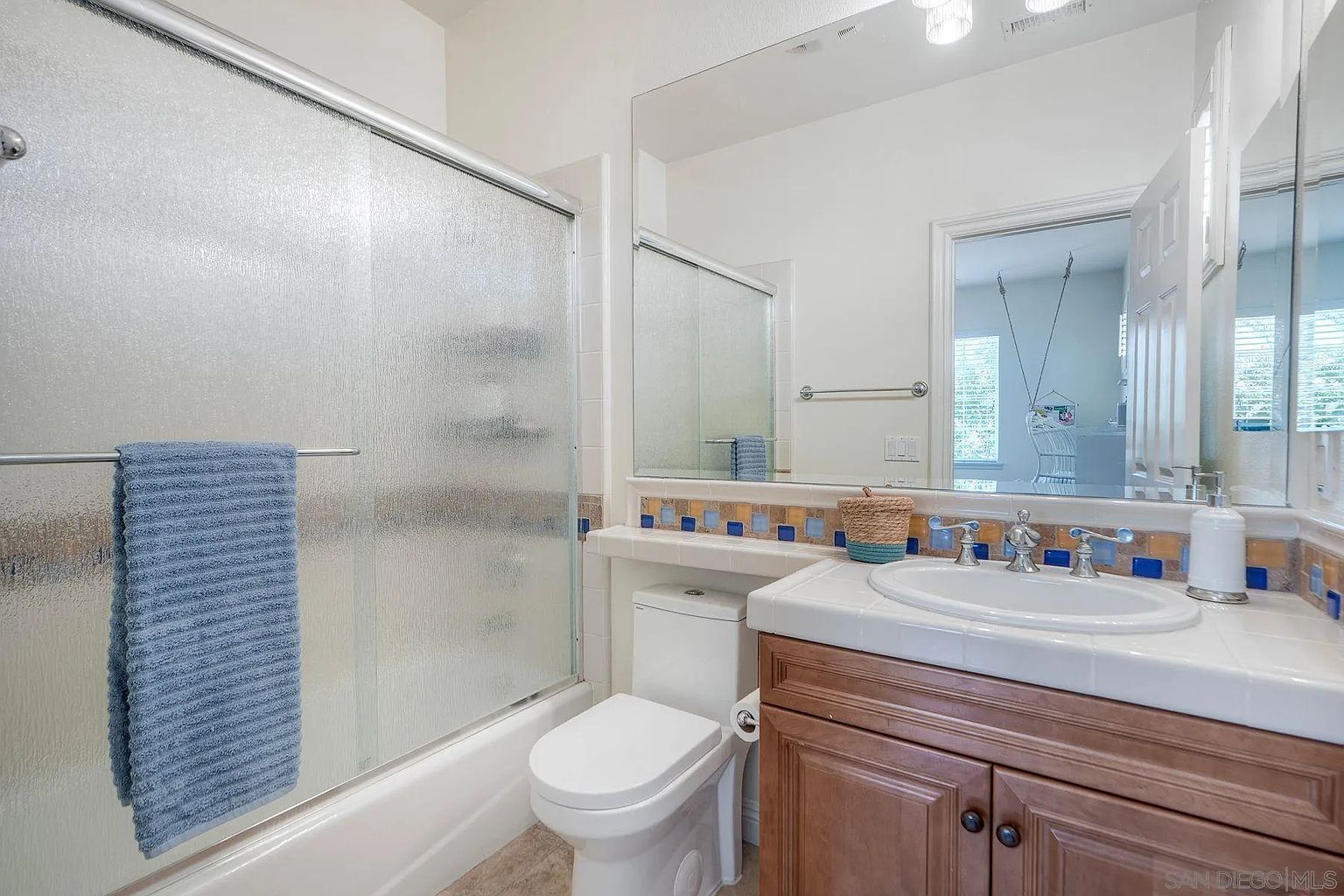 This is a well-maintained guest bathroom featuring a shower-tub combination with a frosted glass enclosure, a white toilet, and a vanity with a white countertop and wooden cabinetry. The mirror above the vanity reflects the bathroom and a glimpse into an adjacent room, enhancing the sense of space. The bathroom has a clean and functional design.