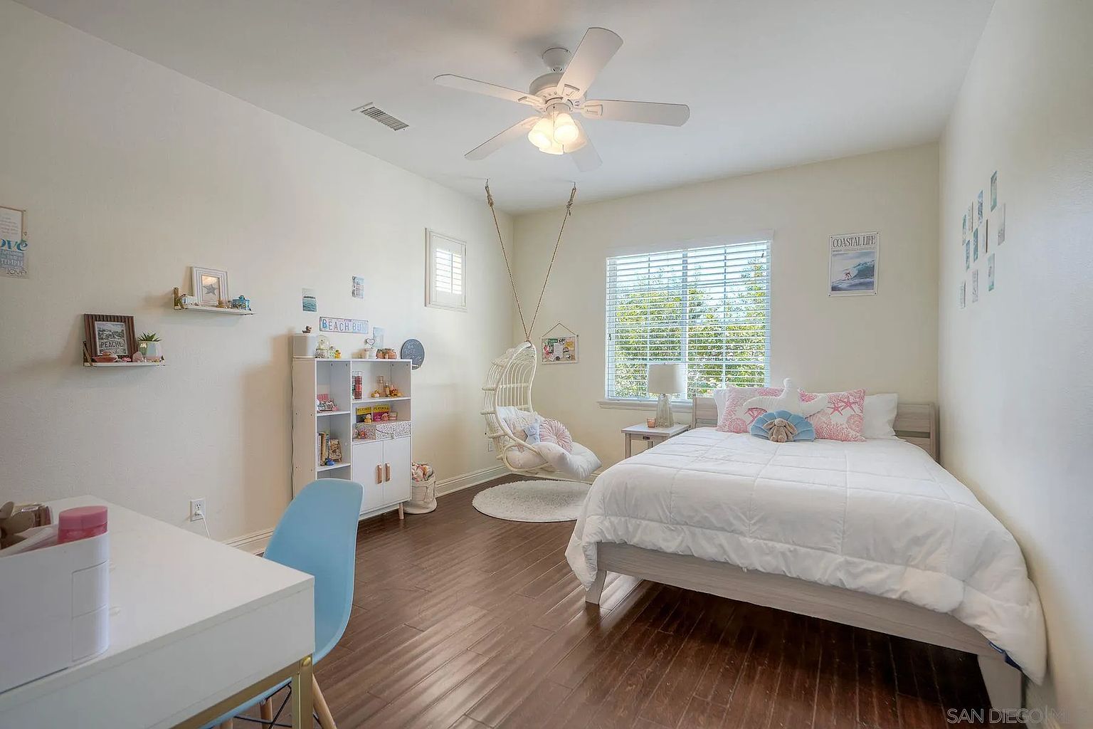 This is a bright and airy bedroom featuring a bed with white bedding, a hanging chair, and a white bookshelf. The room has hardwood floors and a ceiling fan. The overall style is coastal and inviting, perfect for a child or guest room.