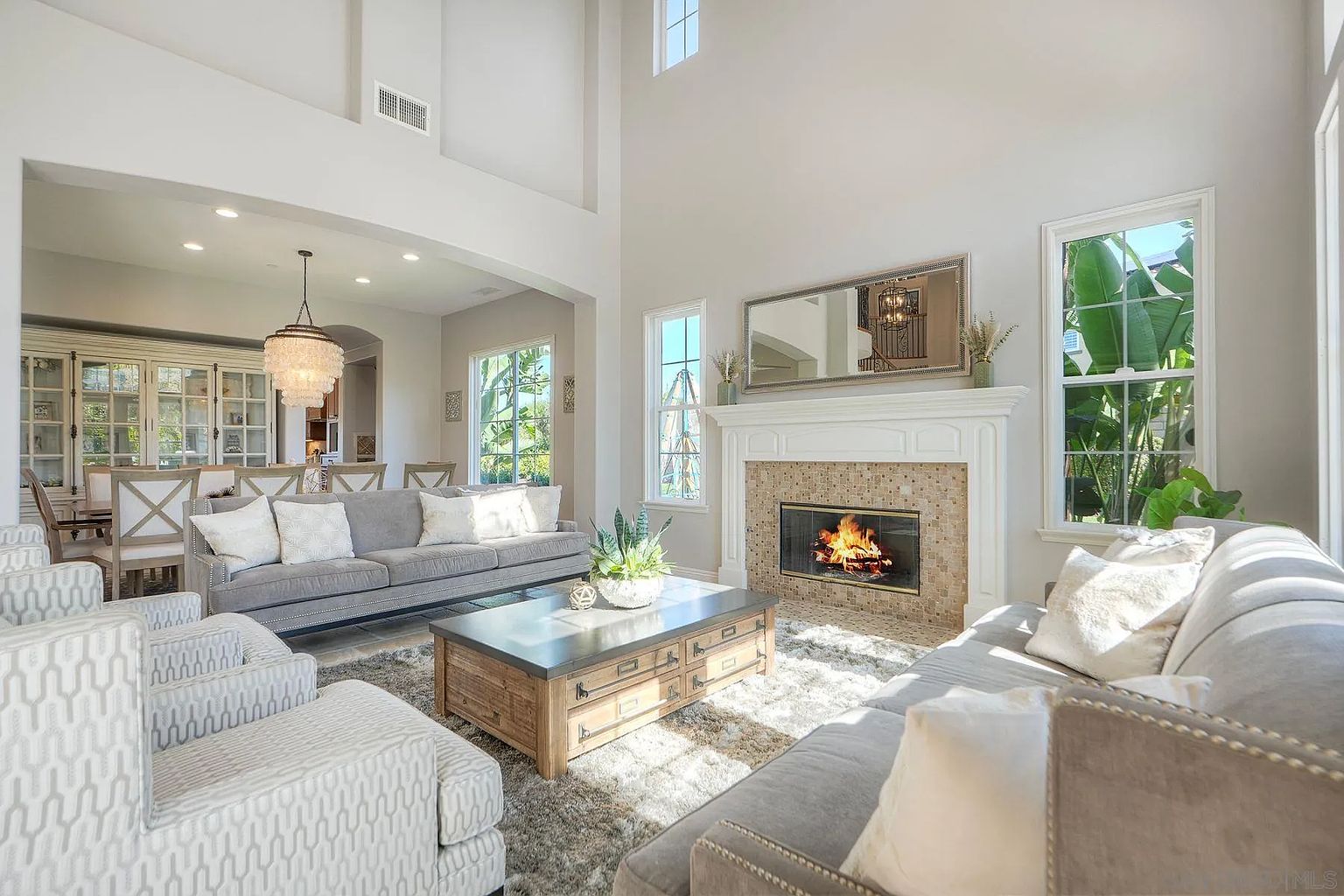 This is a bright and airy living room featuring a high ceiling, a fireplace with a decorative mantel, and large windows that let in plenty of natural light. The room is furnished with comfortable sofas, armchairs, and a coffee table, creating a welcoming and stylish space. The color palette is neutral, with pops of color from the plants and fireplace tiles.