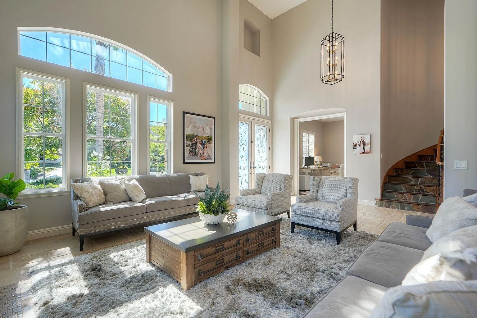 This is a bright and spacious living room featuring high ceilings and large windows that flood the space with natural light. The room is furnished with two sofas, two armchairs, and a rustic coffee table, all arranged on a plush area rug. A staircase is visible on the right side of the image, and a doorway leads to another room, suggesting an open and inviting floor plan.