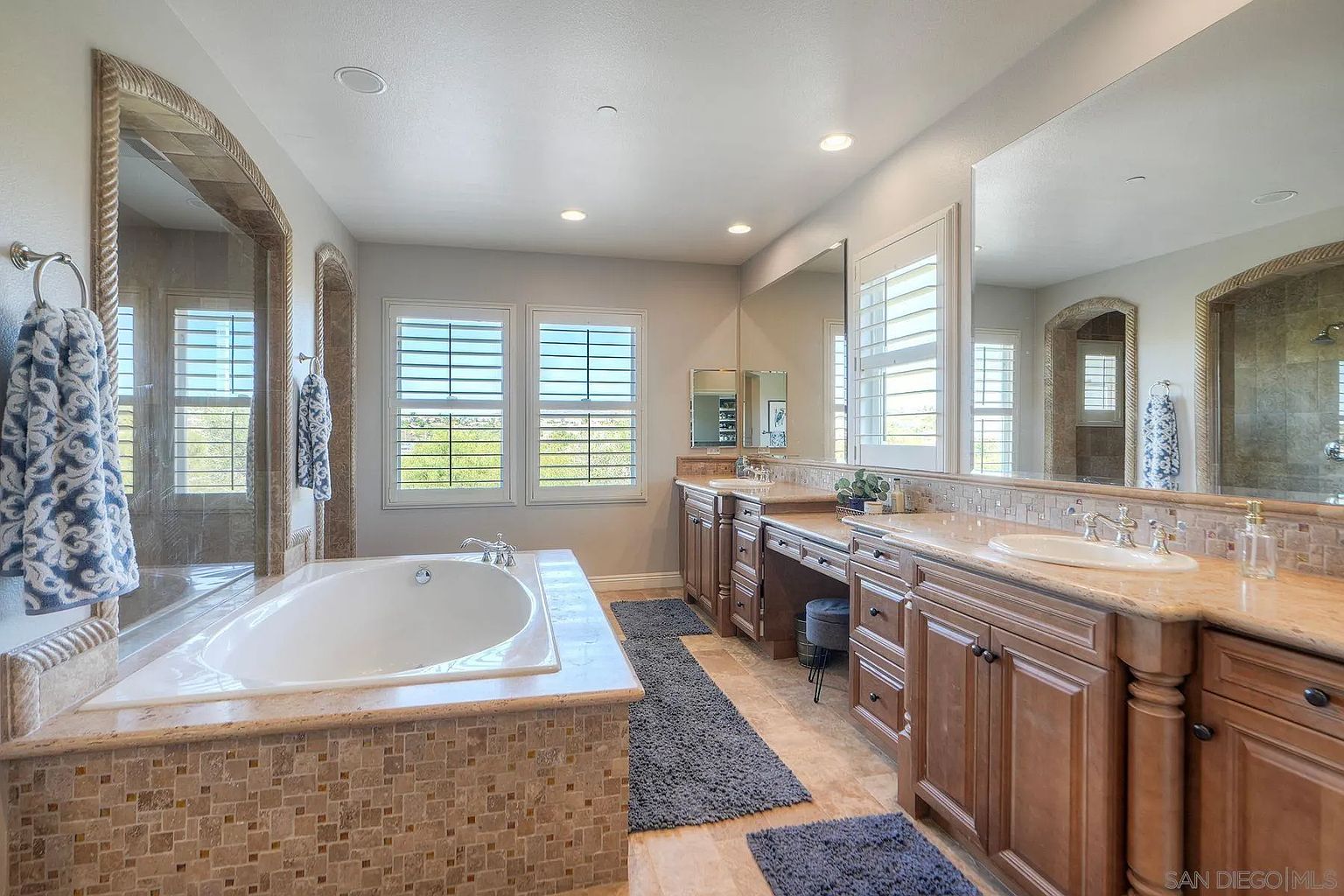 This is a primary bathroom featuring a large soaking tub with a tiled surround, a double vanity with warm-toned countertops and wooden cabinetry, and a large mirror reflecting the space. The bathroom has natural light coming through shuttered windows, and the overall style is luxurious and well-appointed, creating a spa-like atmosphere.