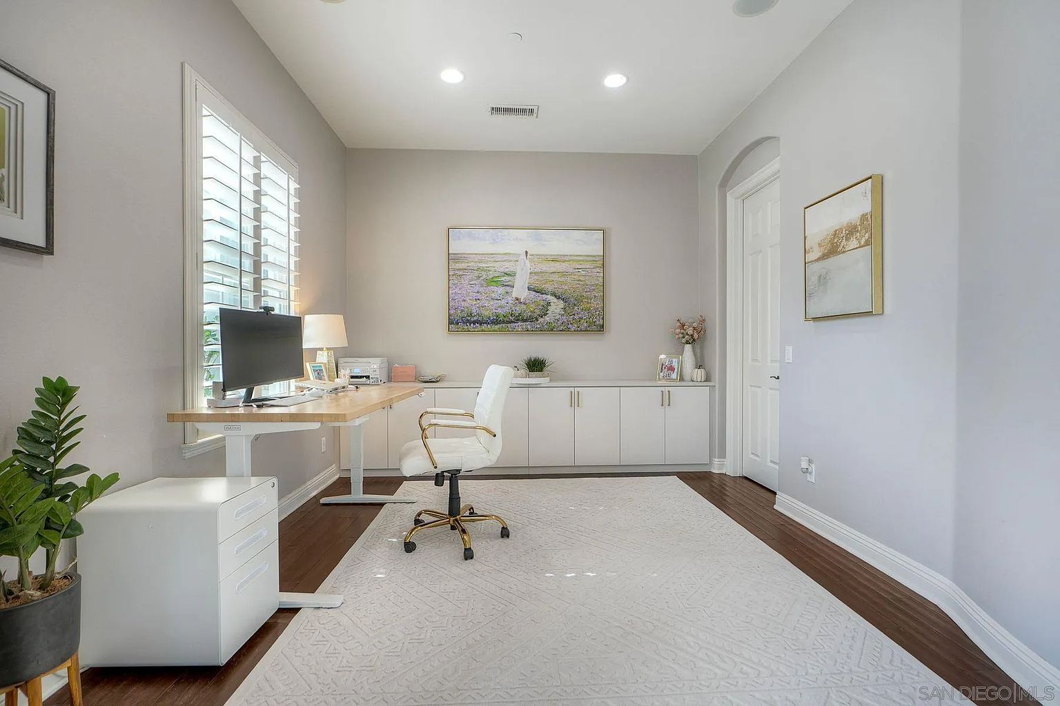 This is an interior shot of a home office featuring a light wood desk with a computer, a white ergonomic chair, and a white filing cabinet. The room is painted in neutral tones, with a large window providing natural light. A built-in cabinet runs along the back wall, and a large area rug covers the dark wood floor, creating a bright and functional workspace.