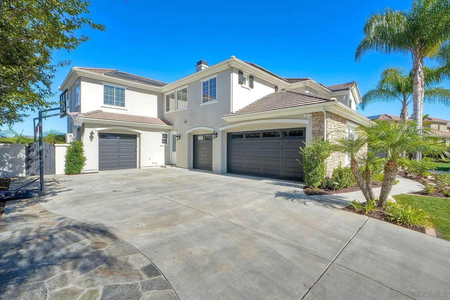 This is a front exterior view of a two-story house with a three-car garage. The house features a combination of stucco and stone accents, a tile roof, and well-maintained landscaping. The driveway is spacious and leads to the garages, enhancing the property's curb appeal.
