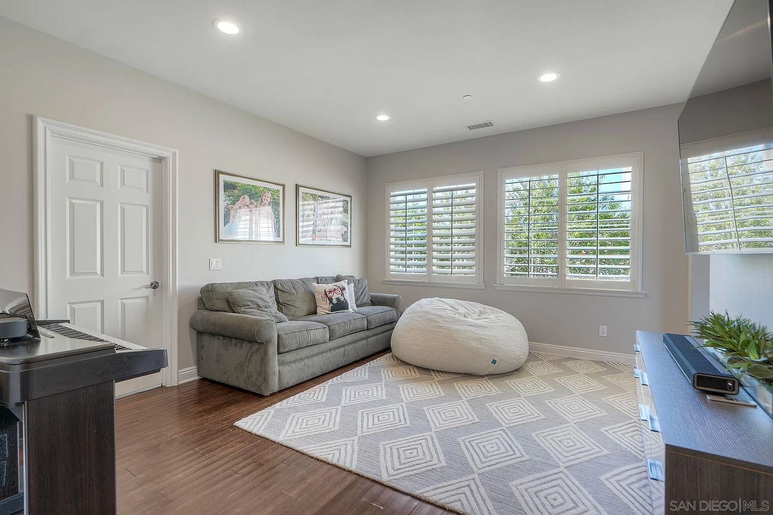 This is an interior shot of a living room featuring a gray sofa with a decorative pillow, a large white beanbag chair, and a gray and white patterned rug. Natural light streams in through shuttered windows, and the room is painted in a neutral gray tone. A piano is visible on the left, and a TV console is on the right, suggesting a comfortable and inviting space for relaxation and entertainment.