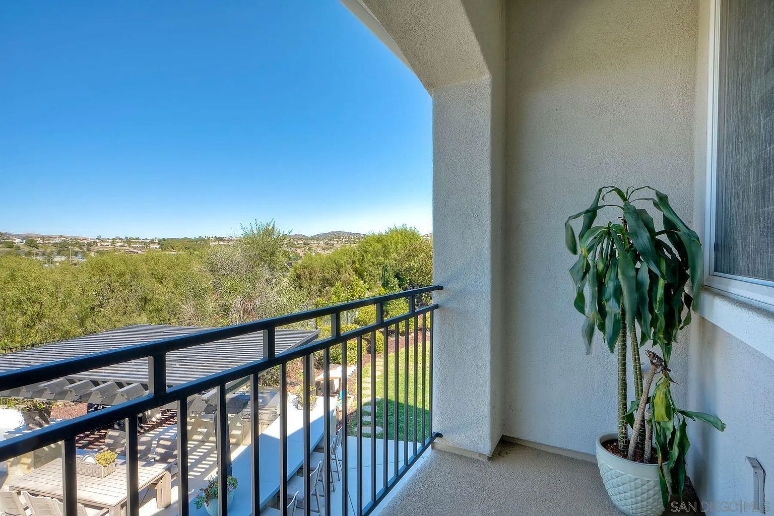 This image showcases a balcony with a black metal railing, offering a scenic view of the surrounding landscape with lush greenery and distant hills under a clear blue sky. A covered patio area with outdoor furniture is visible below, suggesting an inviting space for relaxation and entertainment. A potted plant adds a touch of nature to the balcony space.