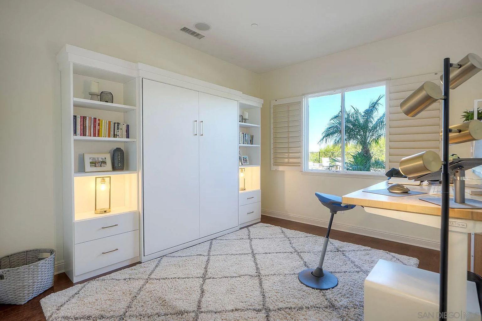 This is an interior shot of a home office featuring a white Murphy bed flanked by built-in bookshelves and drawers. A modern desk with a unique lamp and ergonomic chair sits in front of a window with plantation shutters, offering a view of a palm tree. The room is decorated with a plush, patterned rug, creating a comfortable and functional workspace.