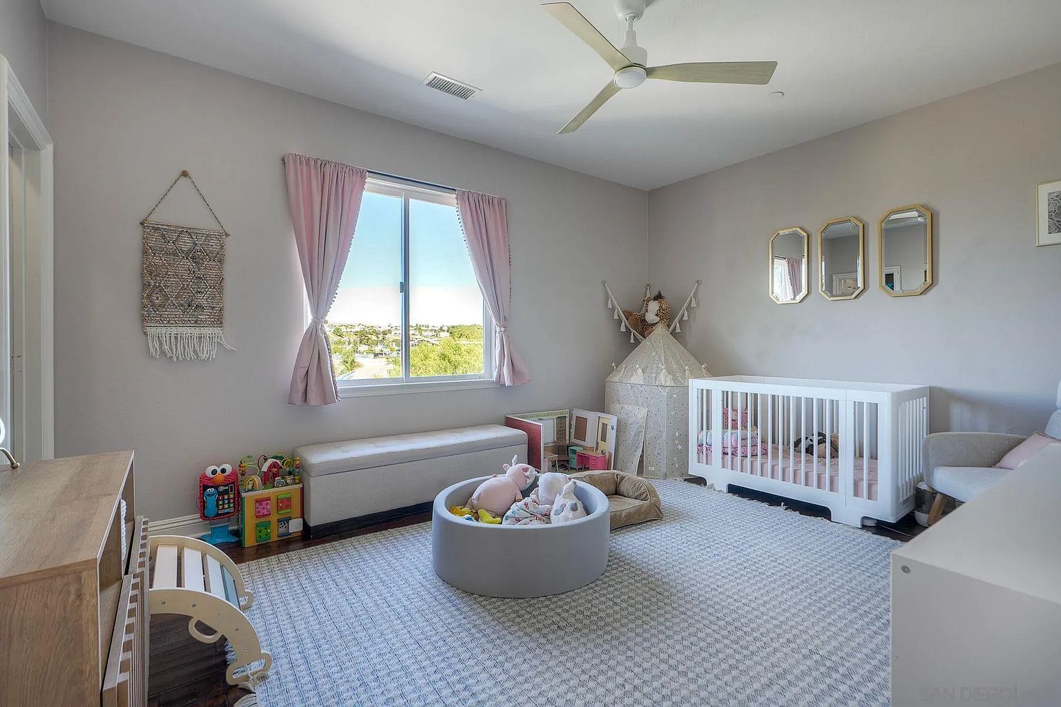 This is a charming nursery featuring a light gray color scheme and a cozy atmosphere. The room includes a white crib, a play tent, a ball pit filled with plush toys, and a window with pink curtains. The overall impression is a comfortable and inviting space for a child.