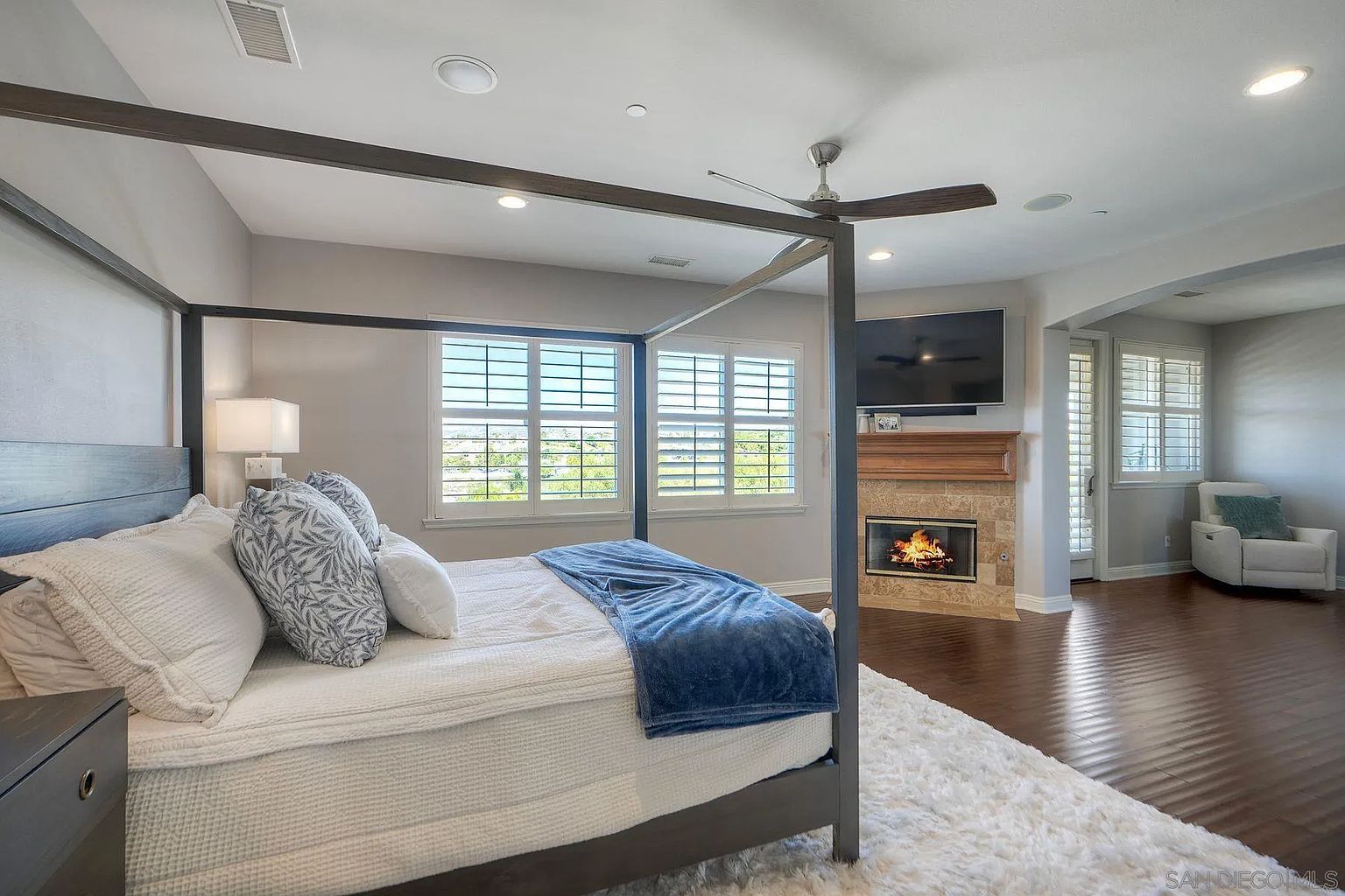 This is a primary bedroom featuring a four-poster bed with white linens and blue accents. The room has a fireplace with a mounted television above it, and large windows with plantation shutters provide natural light. The flooring is dark wood, and a fluffy white rug adds texture to the space.