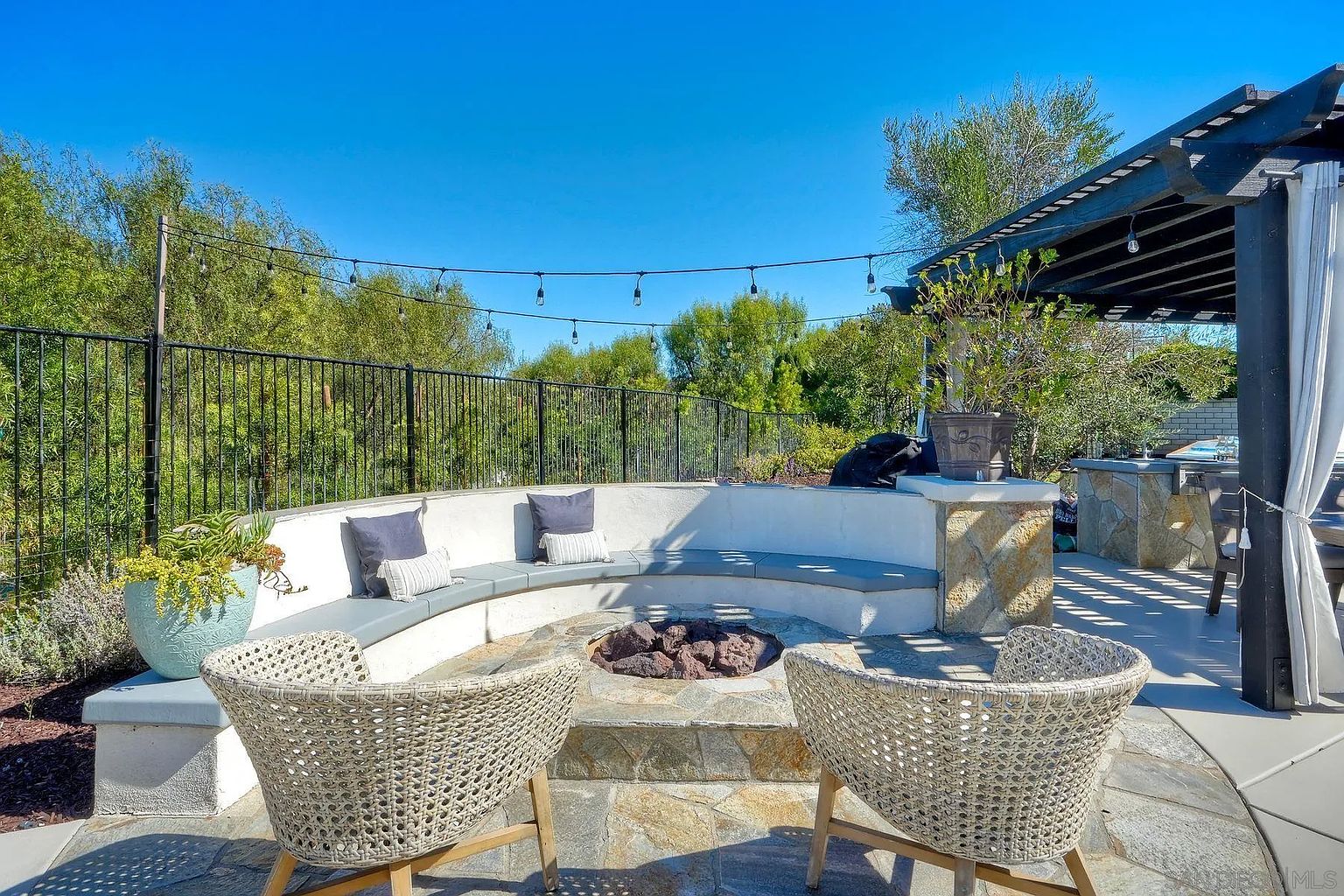 This image showcases a beautifully designed outdoor patio area, featuring a built-in curved seating area with cushions around a central fire pit. Two woven chairs face the fire pit, inviting relaxation and conversation. The space is enhanced by string lights overhead and a pergola structure, creating an inviting and stylish outdoor living space.