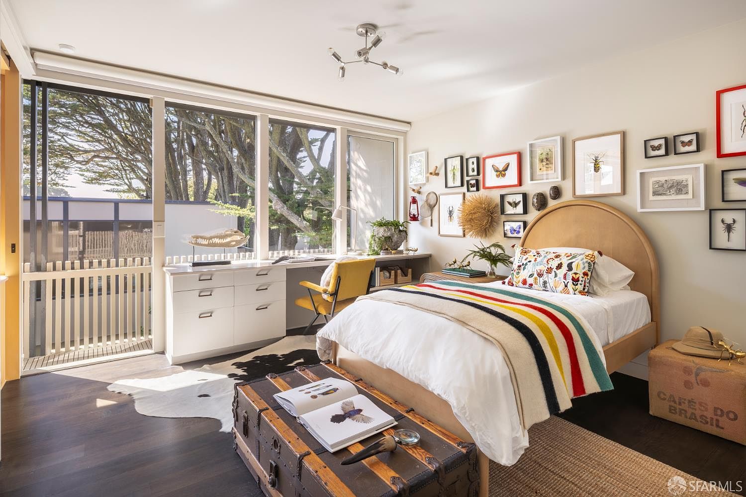 This is a charming bedroom featuring a bed with a colorful striped blanket and decorative pillow, complemented by a gallery wall of framed insects and botanical prints. A desk area is situated by a large window offering natural light and views of the outdoors. The room is decorated with a mix of vintage and modern elements, creating a cozy and inviting atmosphere.