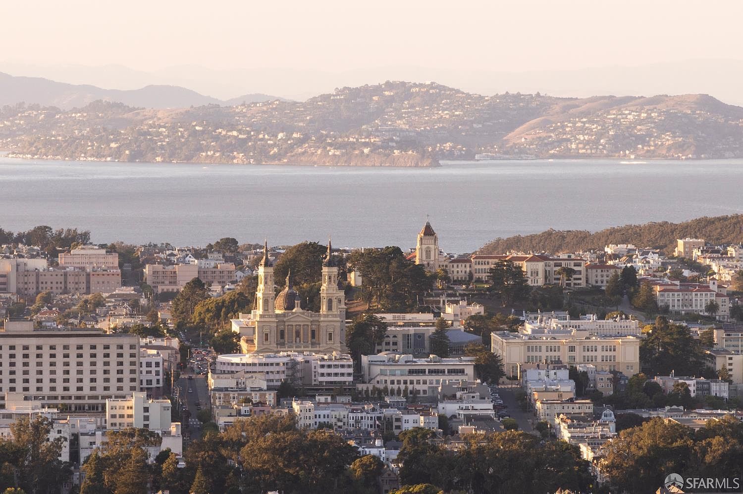 This aerial view showcases a cityscape with prominent buildings, including a church with distinctive domes and a tower. The city extends towards a bay with distant hills in the background, creating a scenic and expansive vista. The overall impression is one of a vibrant urban environment set against a natural backdrop.