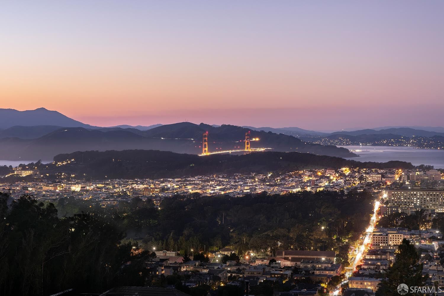 This aerial view captures a cityscape at dusk, featuring the Golden Gate Bridge prominently in the background. The city lights twinkle across the landscape, creating a warm and inviting atmosphere. Mountains are visible in the distance, adding depth to the scene.