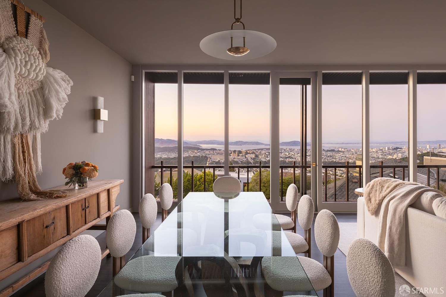 This is an interior shot of a dining room featuring a glass-top table surrounded by modern chairs with textured upholstery. Large windows offer a panoramic view of a cityscape at sunset, creating a sophisticated and inviting atmosphere. A stylish light fixture hangs above the table, and a wooden sideboard with decorative elements adds character to the space.