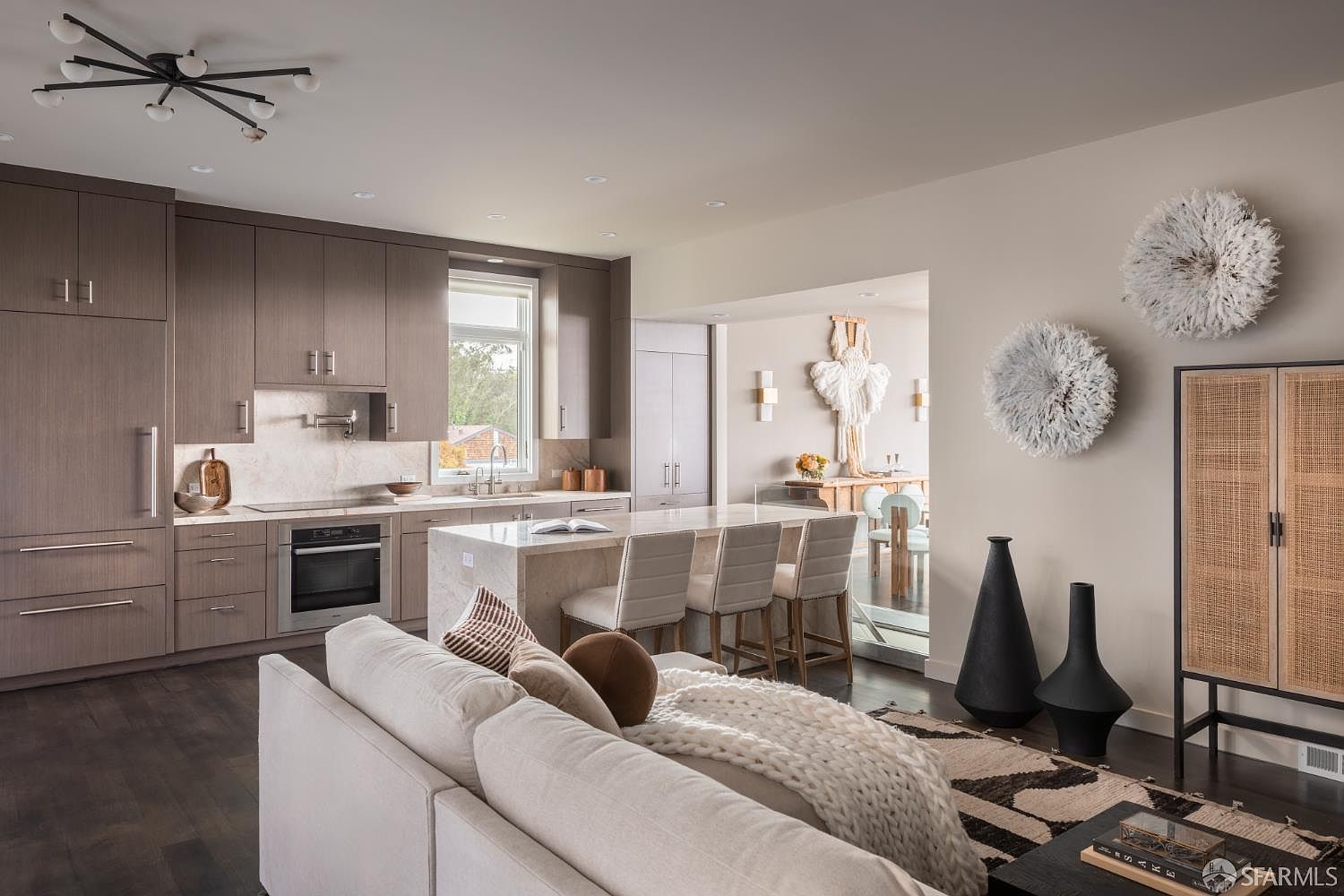 This is an interior shot of a modern living room and kitchen area. The kitchen features sleek, brown cabinetry, stainless steel appliances, and a marble countertop island with bar seating. The living room area includes a comfortable white sofa with decorative pillows and a throw blanket, complemented by black decorative vases and a patterned rug, creating a stylish and inviting space.