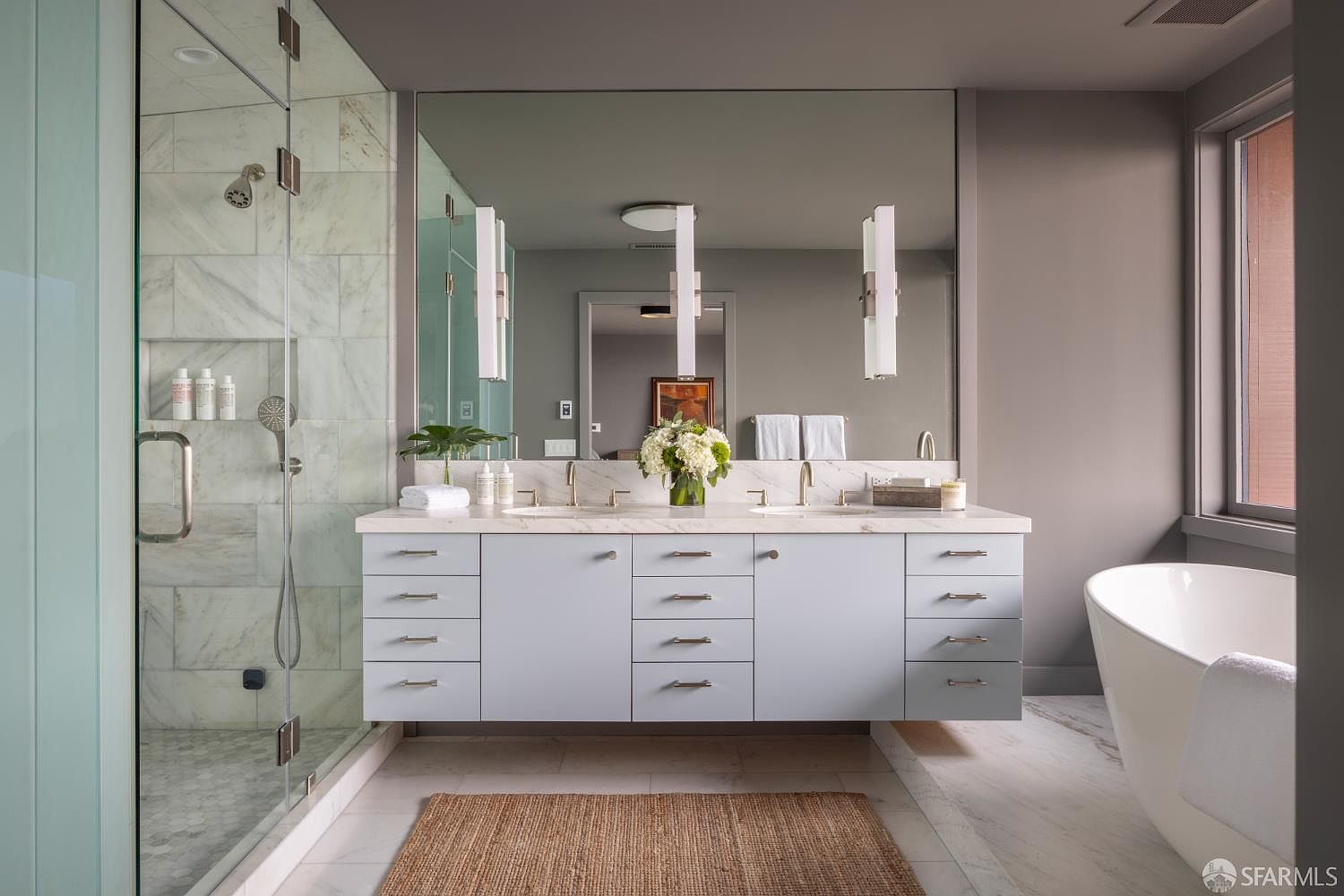 This is a well-lit primary bathroom featuring a double vanity with light blue cabinetry and marble countertops. A large mirror spans the length of the vanity, reflecting pendant lights and a glimpse into another room. A glass-enclosed shower is on the left, and a freestanding bathtub is visible on the right, creating a luxurious and spa-like atmosphere.
