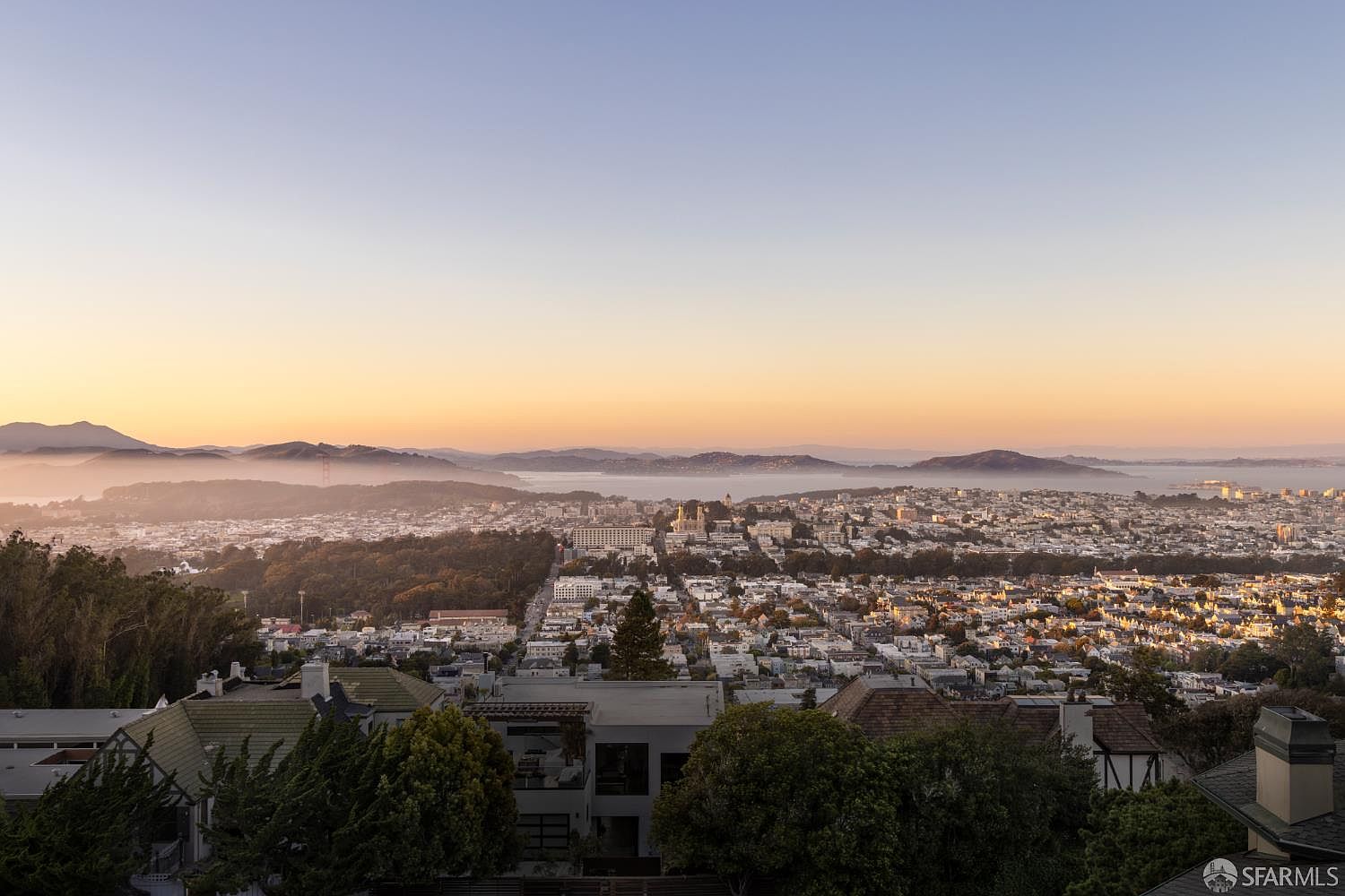 This aerial view showcases a sprawling cityscape at dawn or dusk, with a soft, warm light bathing the buildings and hills in the distance. The city is nestled among rolling hills and a body of water, creating a picturesque scene. The foreground features rooftops and trees, adding depth and perspective to the expansive view.