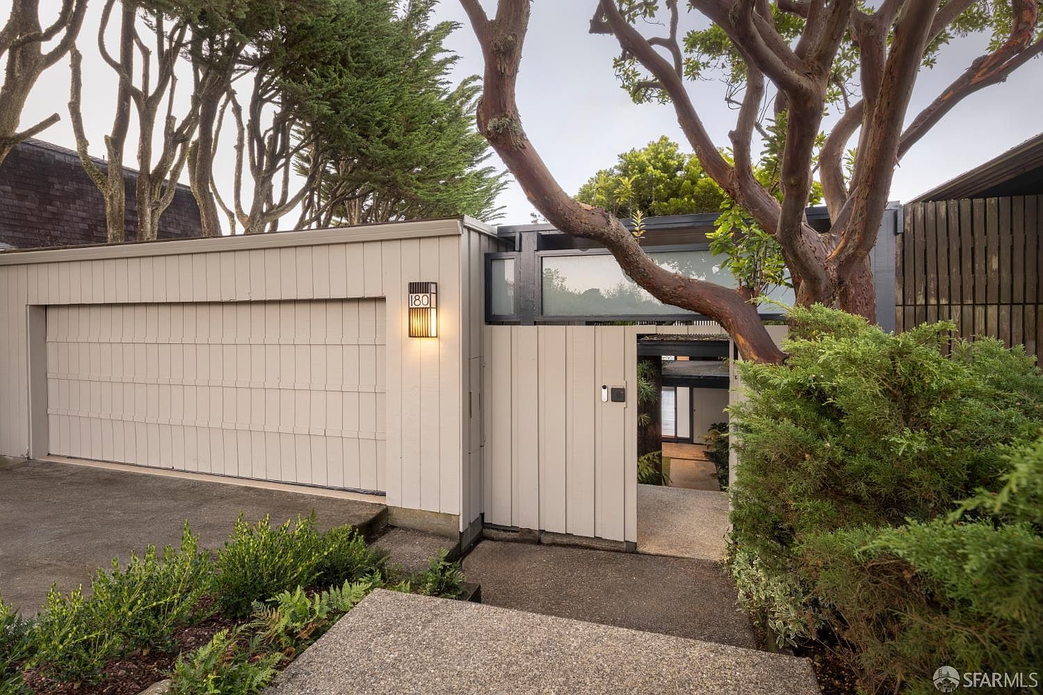 The image showcases the entryway of a modern home, featuring a light-colored garage door and a pedestrian gate. A unique tree with exposed branches adds character to the entrance, while well-maintained greenery enhances the curb appeal. The pathway leads to the front door, creating an inviting and stylish first impression.