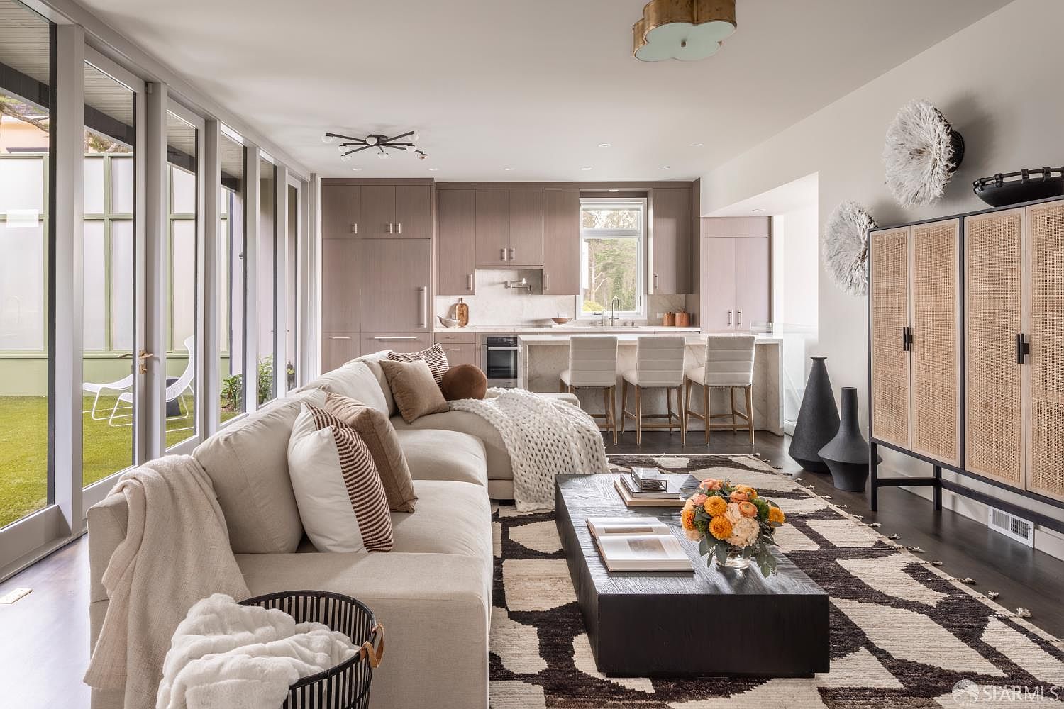 This is an interior shot of a modern living room that seamlessly blends with the kitchen area. The living room features a large, comfortable sectional sofa adorned with throw pillows and blankets, a dark wood coffee table with books and flowers, and a patterned rug. The kitchen is visible in the background with a kitchen island and bar stools, creating an open and inviting space.