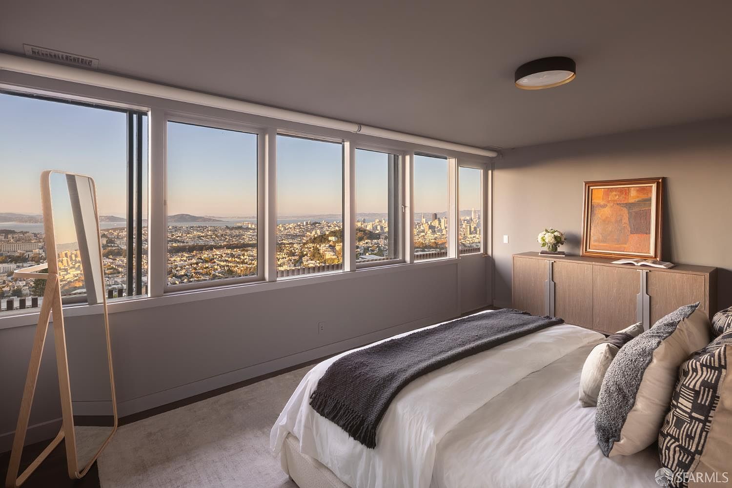 This is a bedroom with a stunning city view through a series of large windows. The room features a bed with white linens and a gray throw, a wooden dresser with artwork above, and a full-length mirror. The color palette is neutral, creating a serene and sophisticated atmosphere.