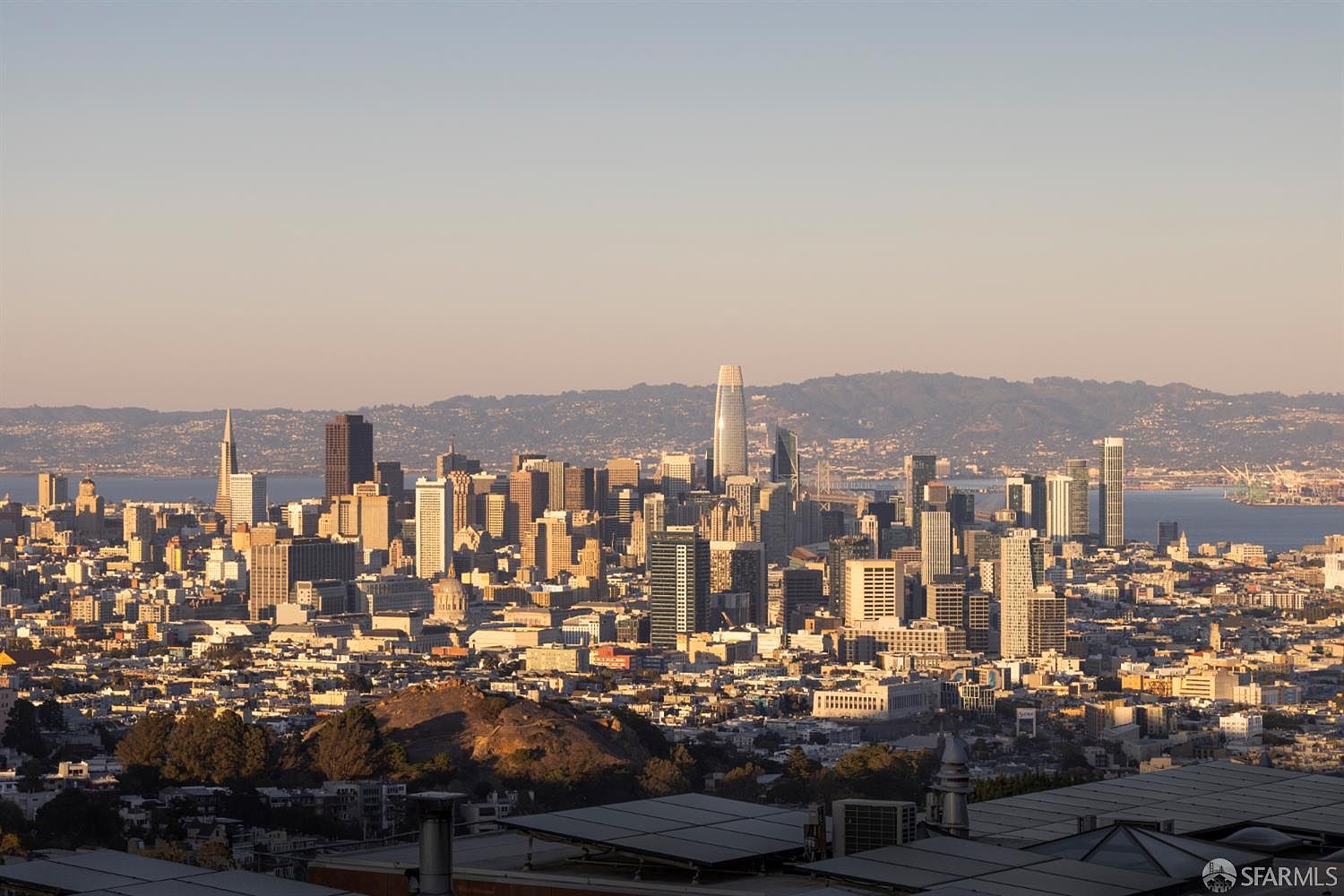 This aerial view showcases the San Francisco skyline, dominated by modern skyscrapers and iconic landmarks. The cityscape is bathed in warm sunlight, highlighting the density of buildings and the surrounding bay. In the foreground, rooftops with solar panels and lush greenery add a touch of urban sustainability.