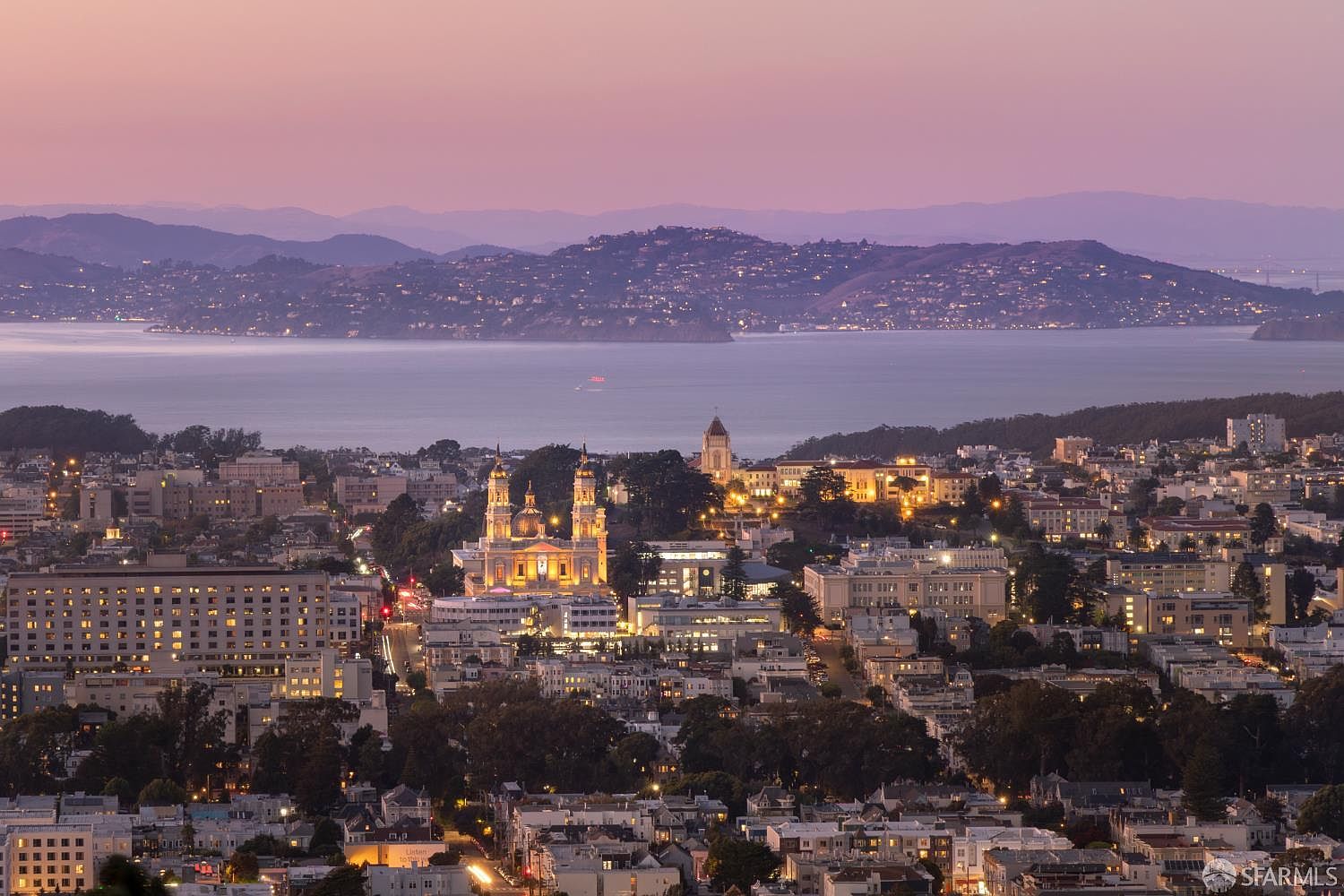 This aerial view showcases a cityscape at dusk, with illuminated buildings and a body of water in the background. The image captures the urban landscape with a focus on the architectural details of the buildings and the soft glow of the city lights. The overall impression is one of a vibrant and thriving urban center.