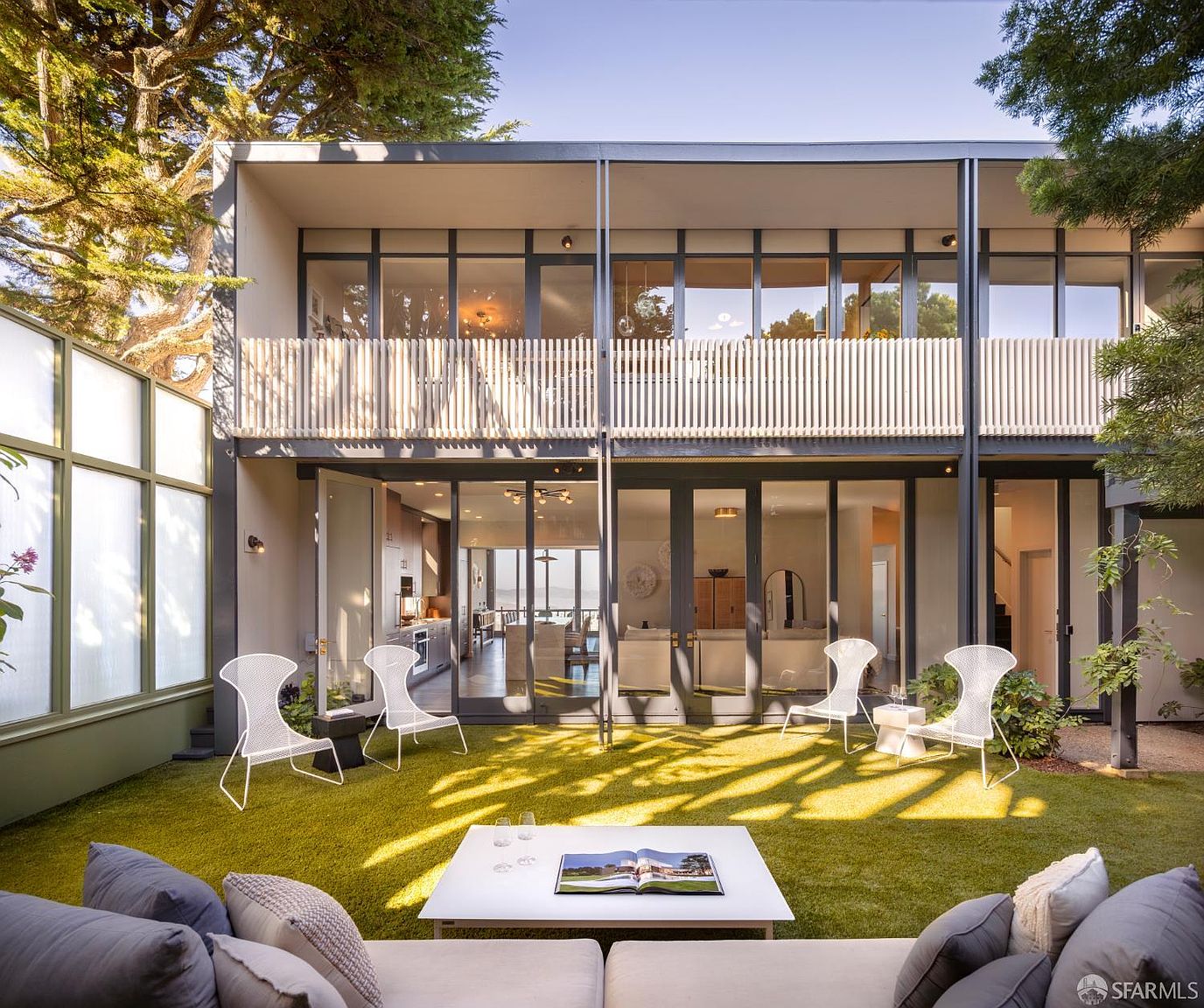 This image showcases the rear exterior of a modern home, emphasizing the yard and garden area. The space features a well-manicured lawn, contemporary outdoor furniture, and large glass doors that seamlessly blend indoor and outdoor living. A balcony with a slatted railing adds architectural interest to the upper level.