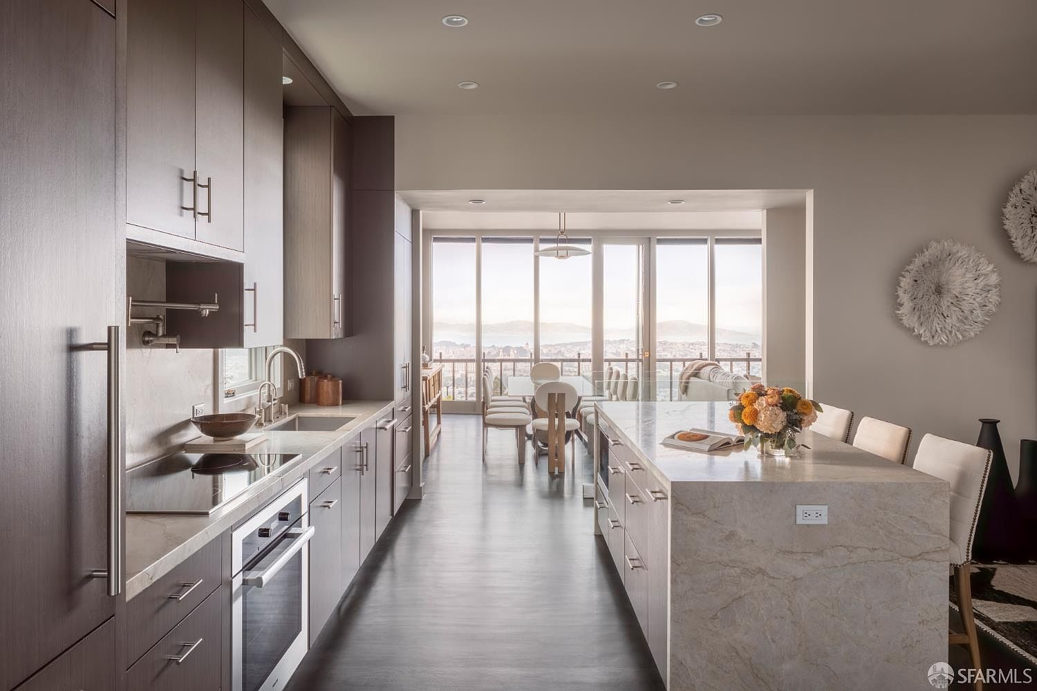 This is a modern kitchen with dark wood cabinetry and a large island with a marble countertop. The kitchen opens to a dining area with floor-to-ceiling windows offering a city view. The space is well-lit and features clean lines, creating a sophisticated and inviting atmosphere.
