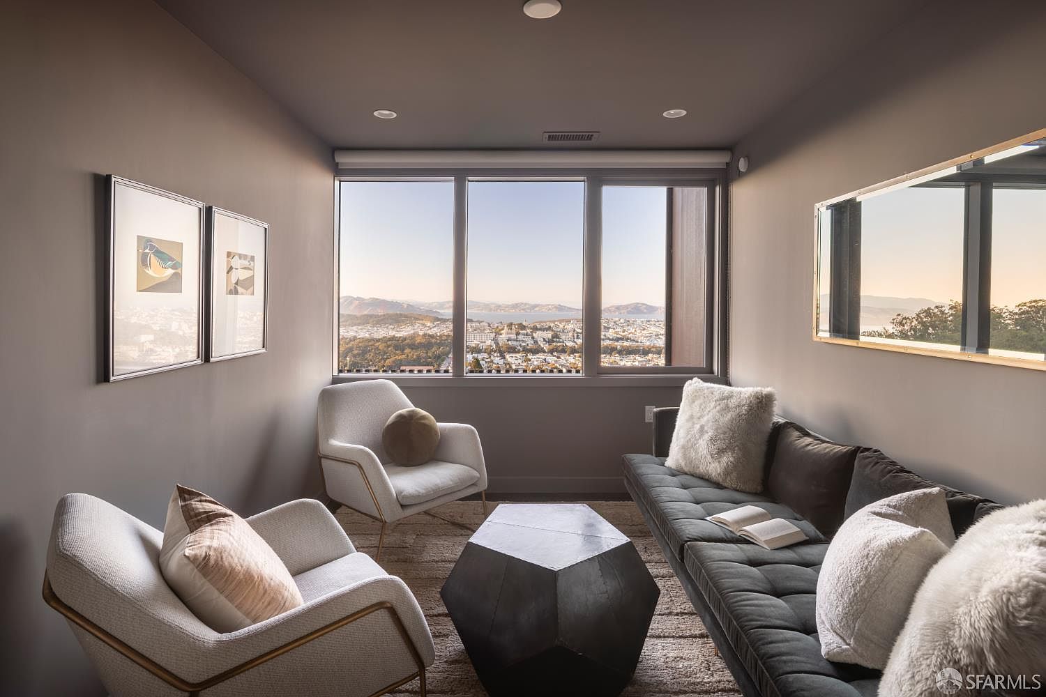 This is an interior shot of a living room featuring two armchairs, a sofa, and a geometric coffee table. The room has a modern aesthetic with neutral tones and artwork on the walls. A large window provides a view of the city and mountains, enhancing the room's appeal.