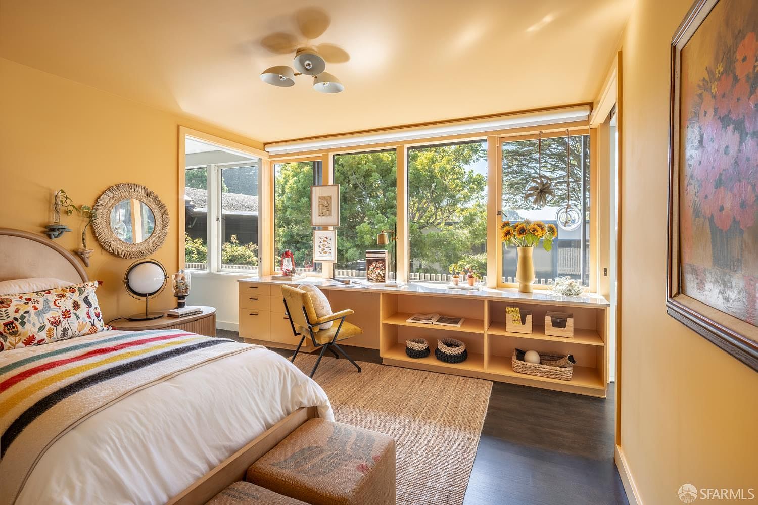 This is a bright and inviting bedroom featuring a large window with a built-in desk and shelving, providing ample natural light and workspace. The room is decorated in warm tones, with a comfortable bed and stylish furnishings, creating a cozy and functional space. The dark wood floors contrast nicely with the light colored walls and furniture.