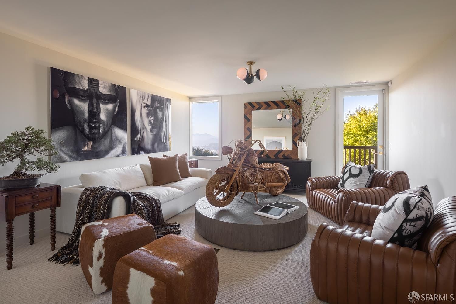 This is an interior shot of a stylish living room featuring a white sofa adorned with brown pillows and a throw blanket. Two brown and white ottomans sit in front of the sofa. The room also includes two brown leather armchairs with patterned pillows, a round coffee table with a decorative motorcycle, and artwork on the wall. The overall aesthetic is modern and sophisticated, with a touch of rustic charm.