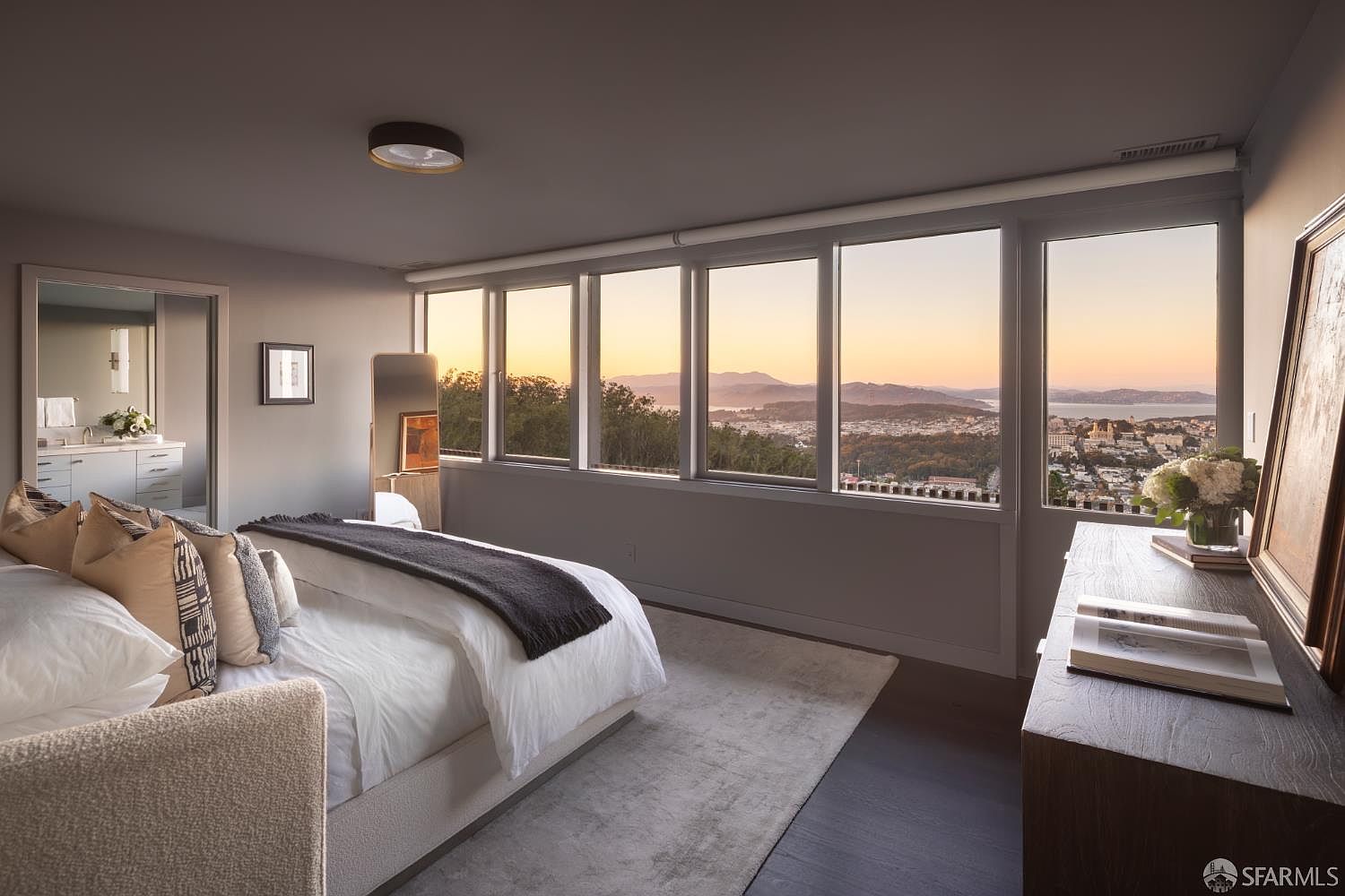 This is a primary bedroom featuring a large bed with multiple pillows and a dark throw blanket. The room has a modern aesthetic with gray walls and dark wood floors, complemented by a light gray rug. Large windows offer a stunning view of the city and surrounding landscape, enhancing the room's appeal.