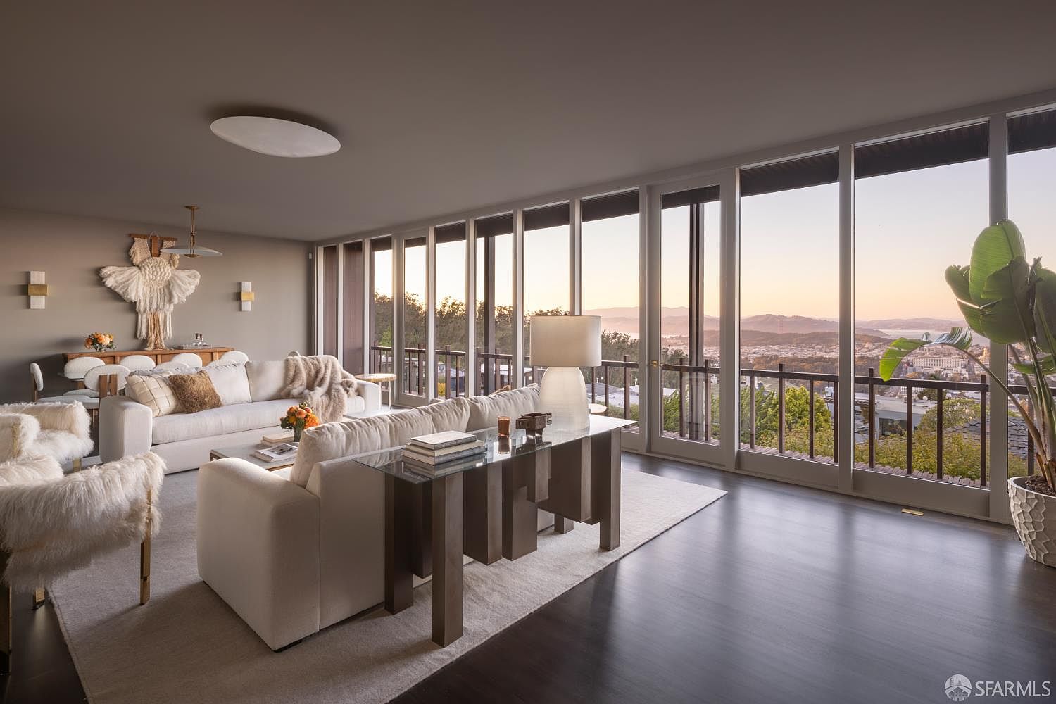 This is an interior shot of a luxurious living room featuring modern design elements. The room is furnished with white sofas, a glass-topped console table, and a decorative wall hanging. Large windows offer a stunning view of the city and bay, enhancing the room's appeal and creating a bright, airy atmosphere.