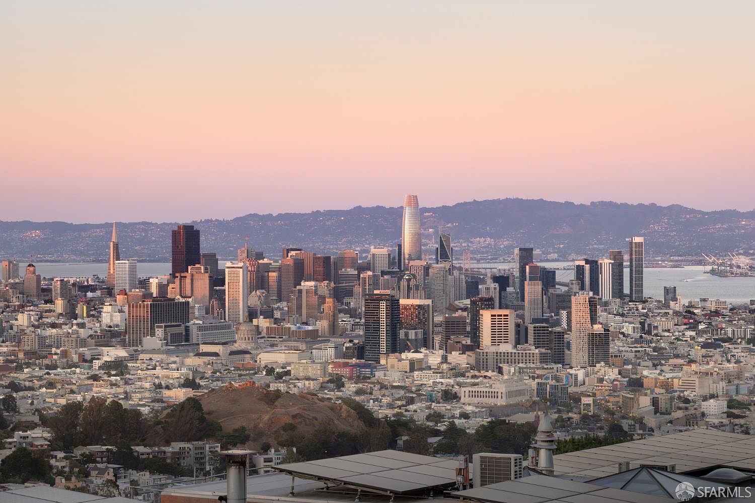 This aerial view showcases the San Francisco skyline at dusk, with prominent skyscrapers and a soft, pastel-colored sky. The city's layout is visible, extending towards the bay and distant hills. Solar panels are visible in the foreground, suggesting a focus on sustainability.