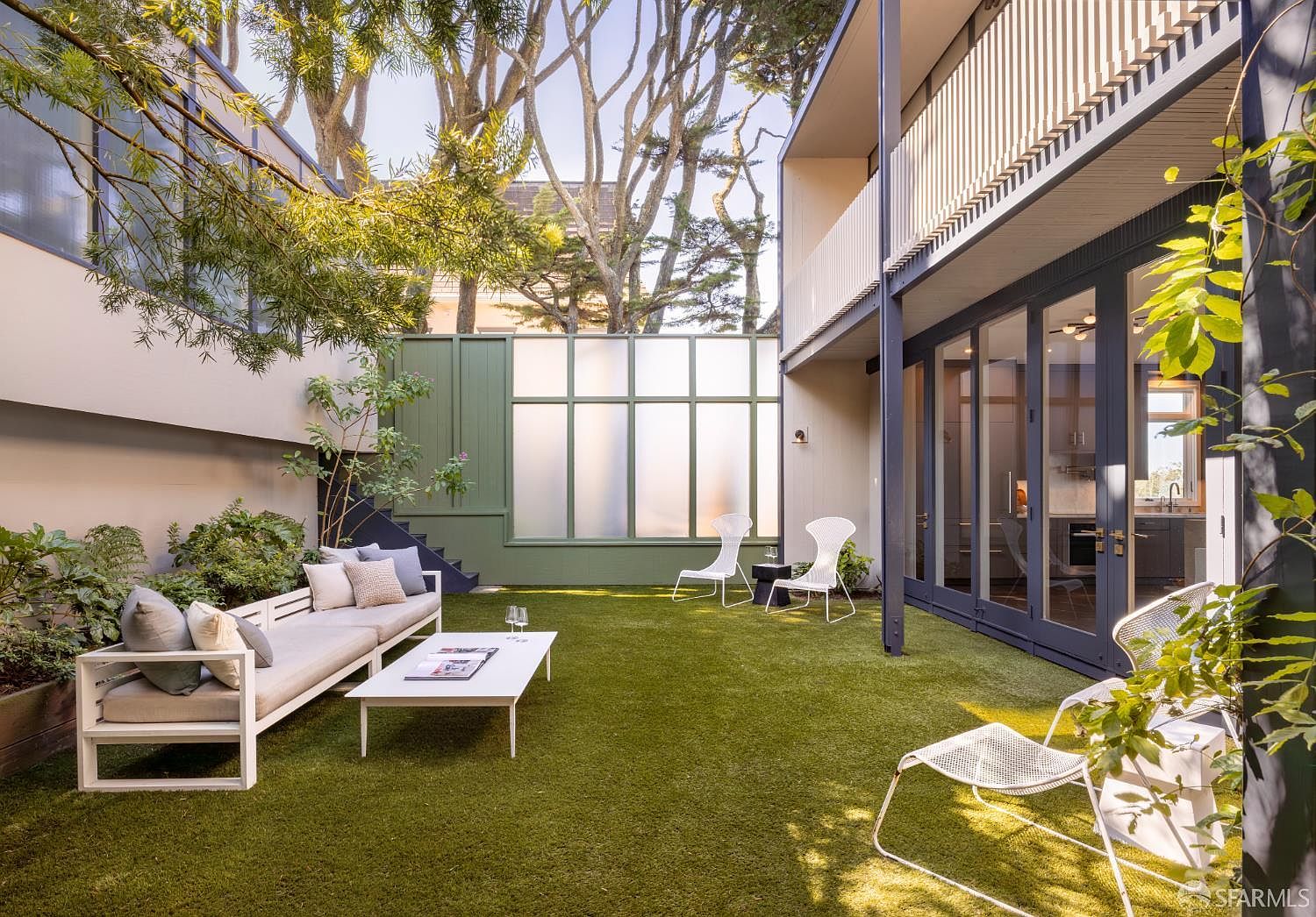 This image showcases a modern backyard garden area. It features a comfortable outdoor seating arrangement with a sofa and coffee table on artificial turf. The space is enclosed by a combination of walls, a frosted glass panel, and lush greenery, creating a private and stylish outdoor living area.