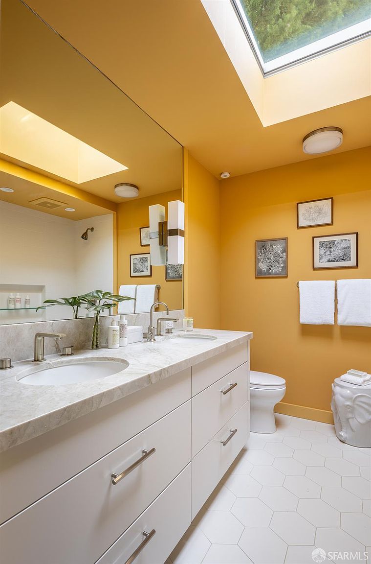 This is a well-lit primary bathroom featuring a double vanity with a white marble countertop and white cabinetry. The walls are painted a warm yellow, and the floor is covered in white hexagonal tiles. A large mirror reflects the vanity and a skylight, enhancing the natural light in the space.