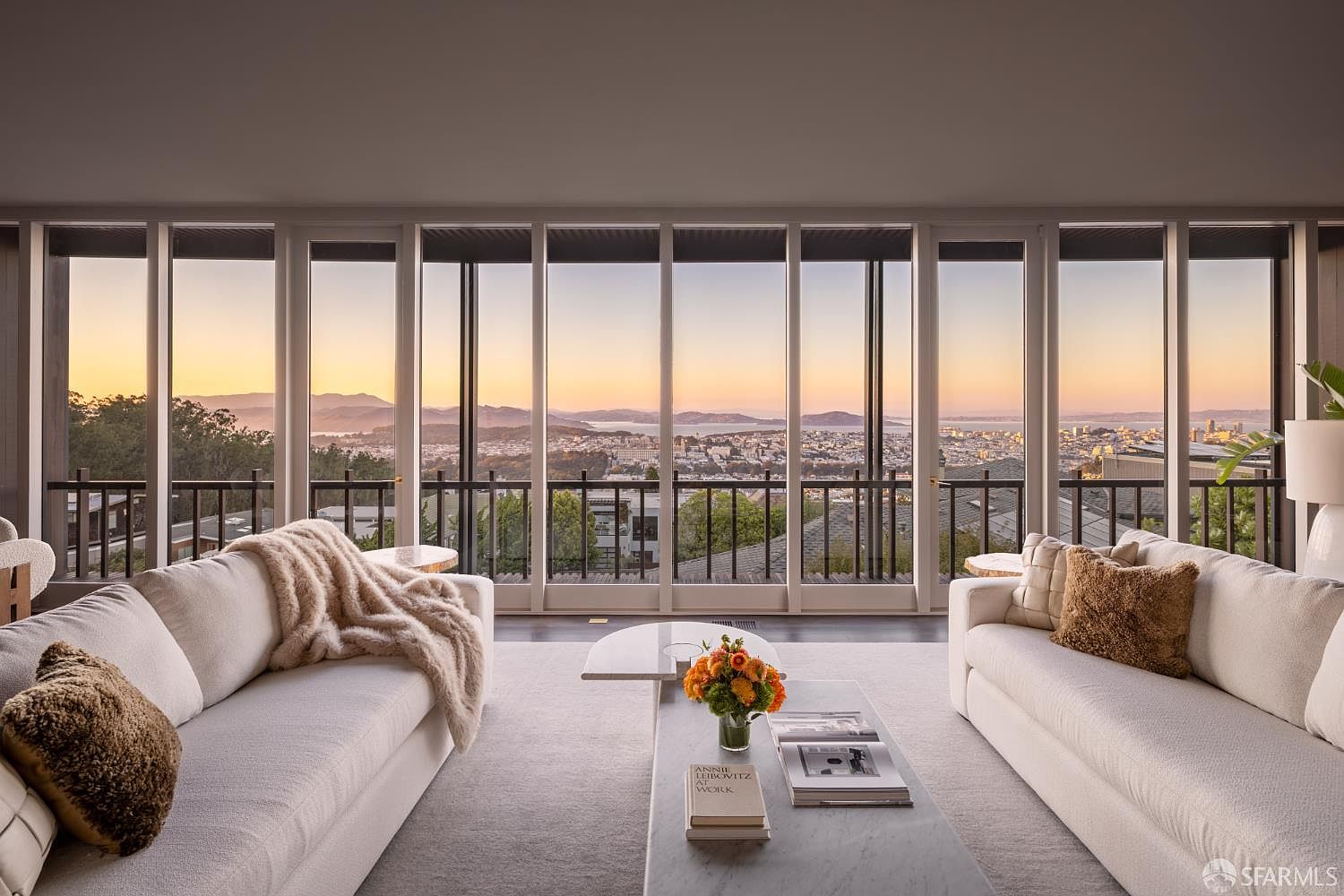 This is a bright and airy living room featuring floor-to-ceiling windows that offer a panoramic view of the city and surrounding landscape. The room is furnished with two white sofas, a marble coffee table with floral arrangement, and neutral-toned pillows and throws, creating a comfortable and elegant space. The overall impression is one of luxury and sophistication, emphasizing the stunning view and natural light.
