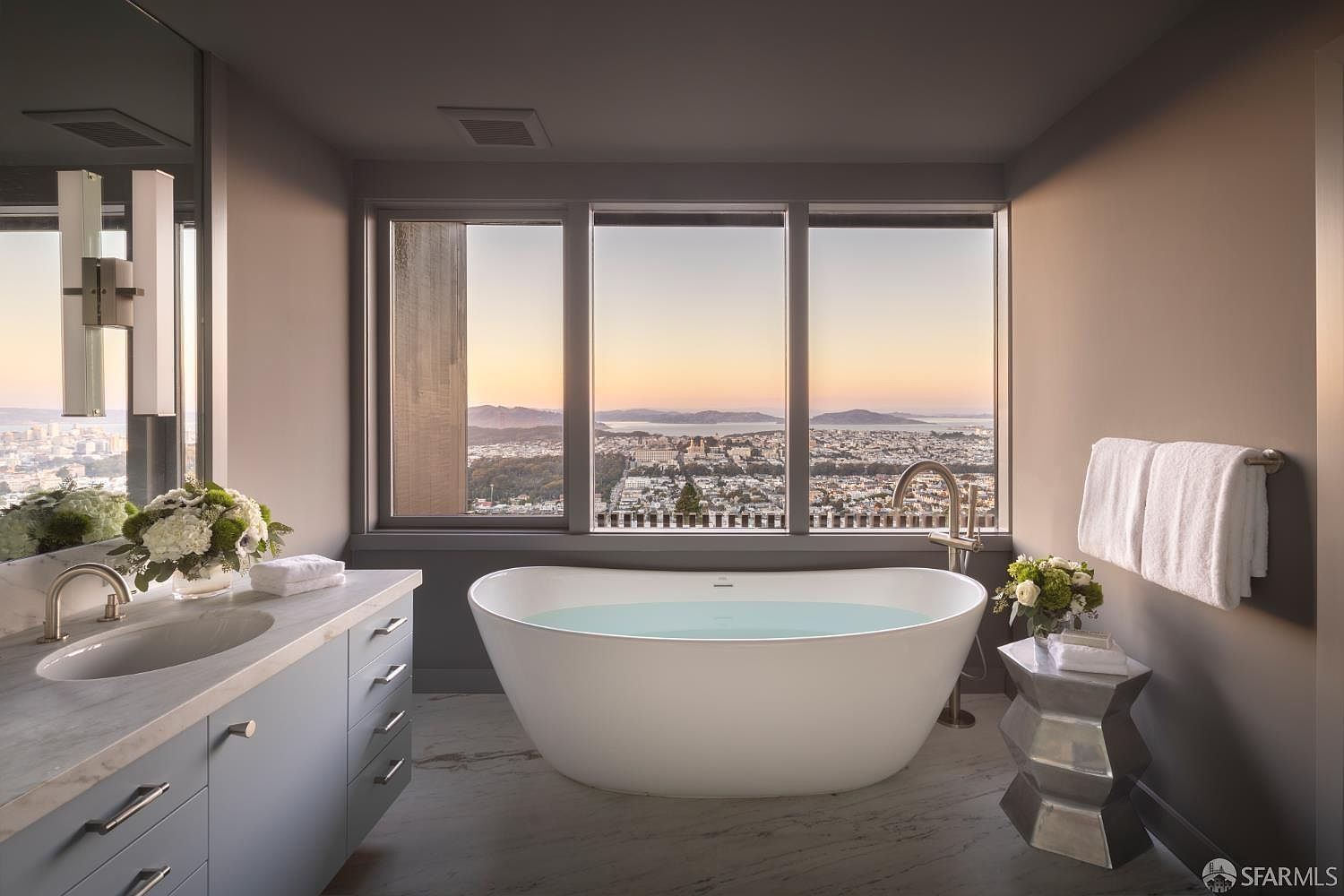 This is a luxurious primary bathroom featuring a freestanding soaking tub positioned in front of a large window with a stunning city view. The bathroom includes a vanity with a marble countertop, modern fixtures, and a neutral color palette, creating a serene and upscale atmosphere. The perspective is from the doorway, showcasing the tub as the focal point.