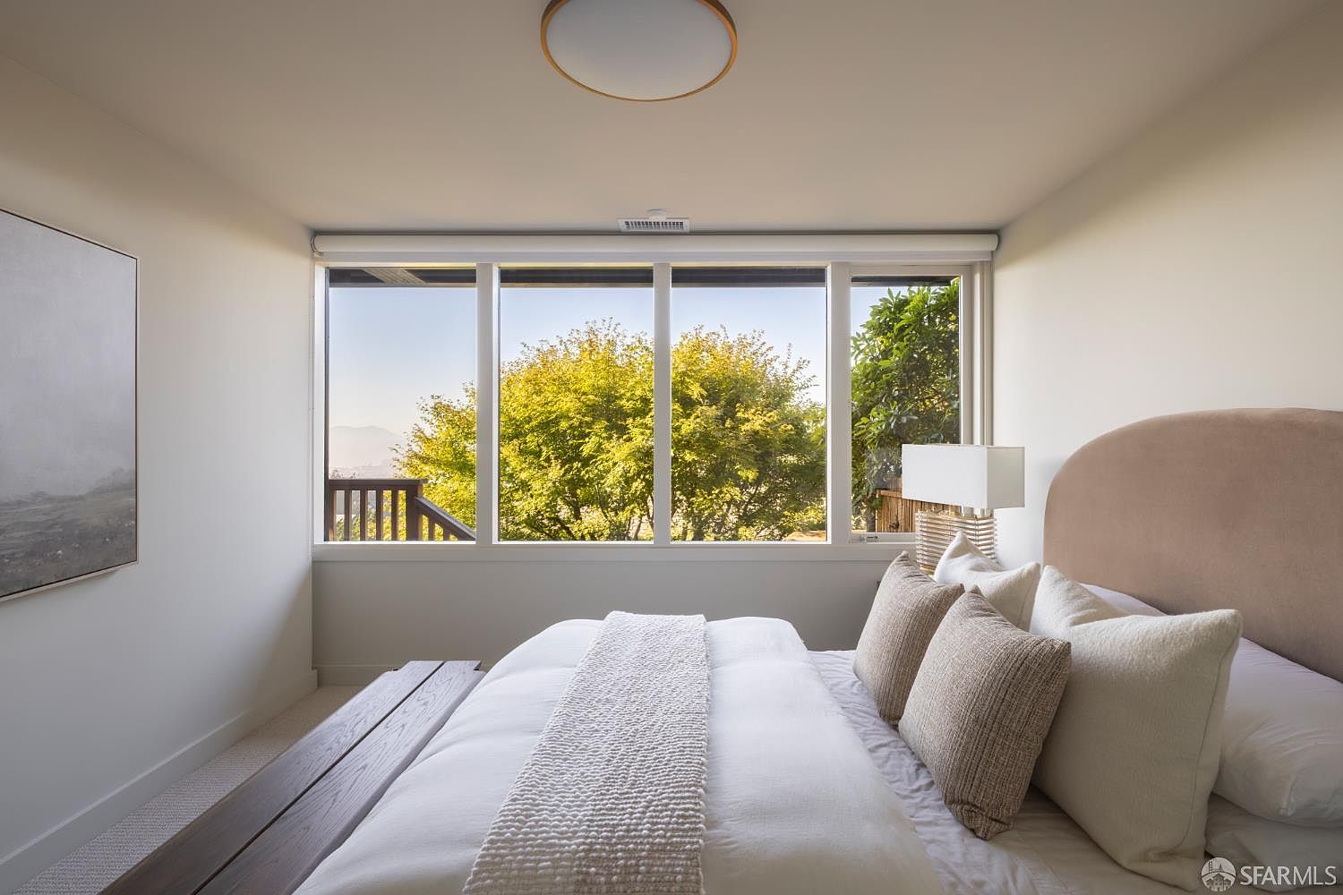 This is a bright and airy primary bedroom featuring a large window with a view of lush greenery. The room is decorated in neutral tones with a light-colored bedspread and pillows, complemented by a padded headboard. A modern ceiling light fixture and artwork add subtle sophistication to the space.